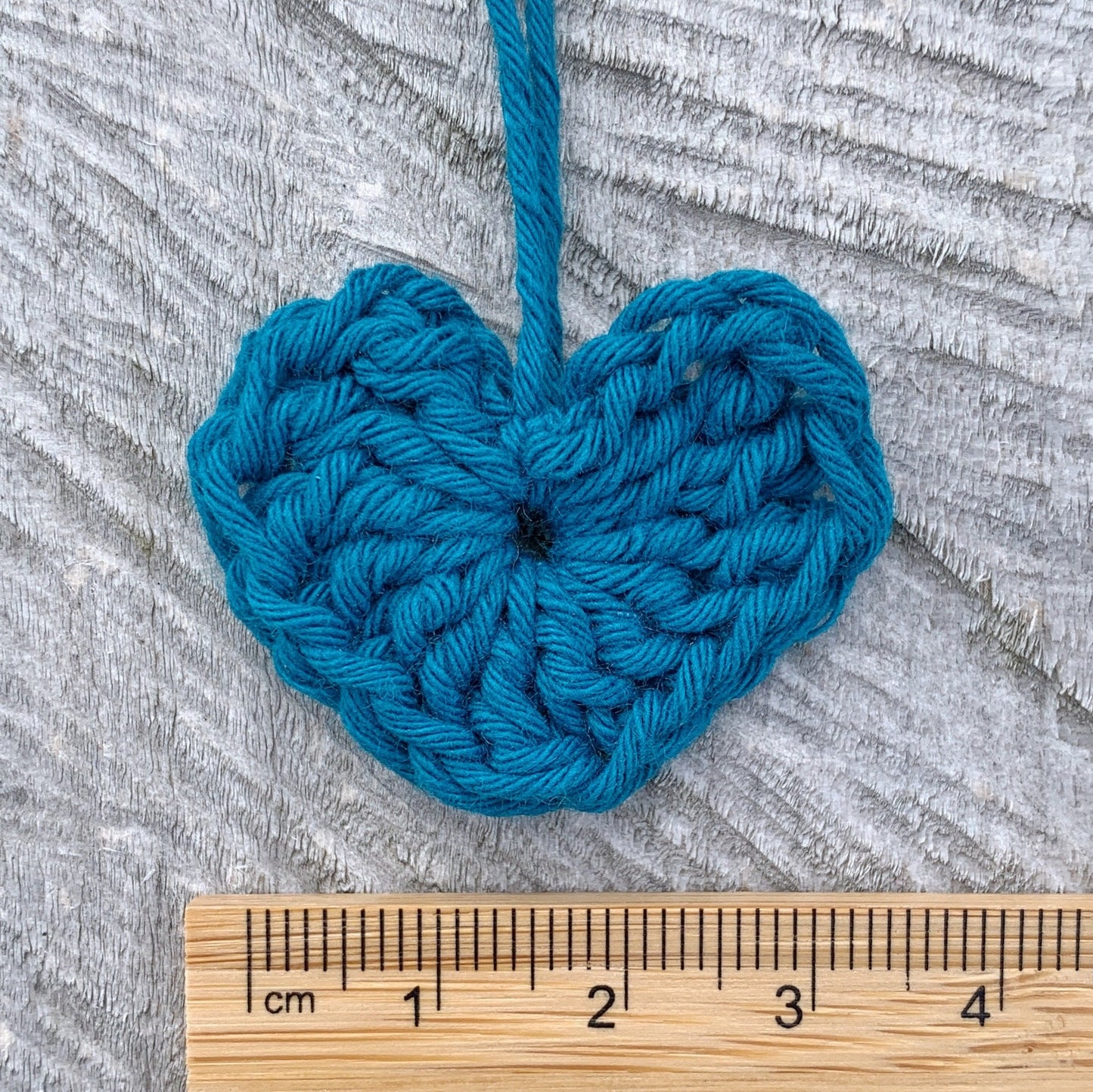 Teal crocheted heart with a wooden ruler for scale on a textured grey background