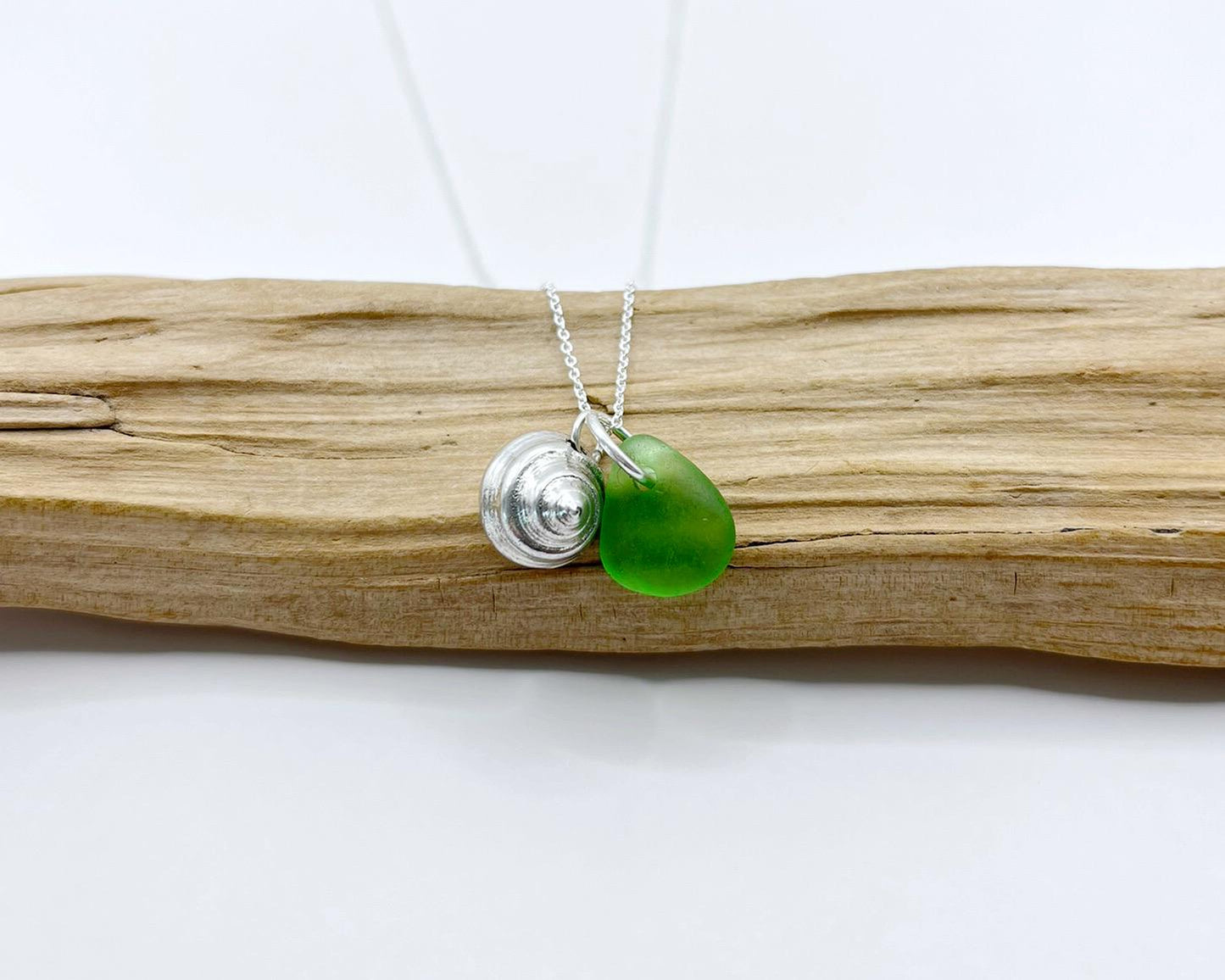 Necklace with a silver shell and green sea glass laid on driftwood against a white background