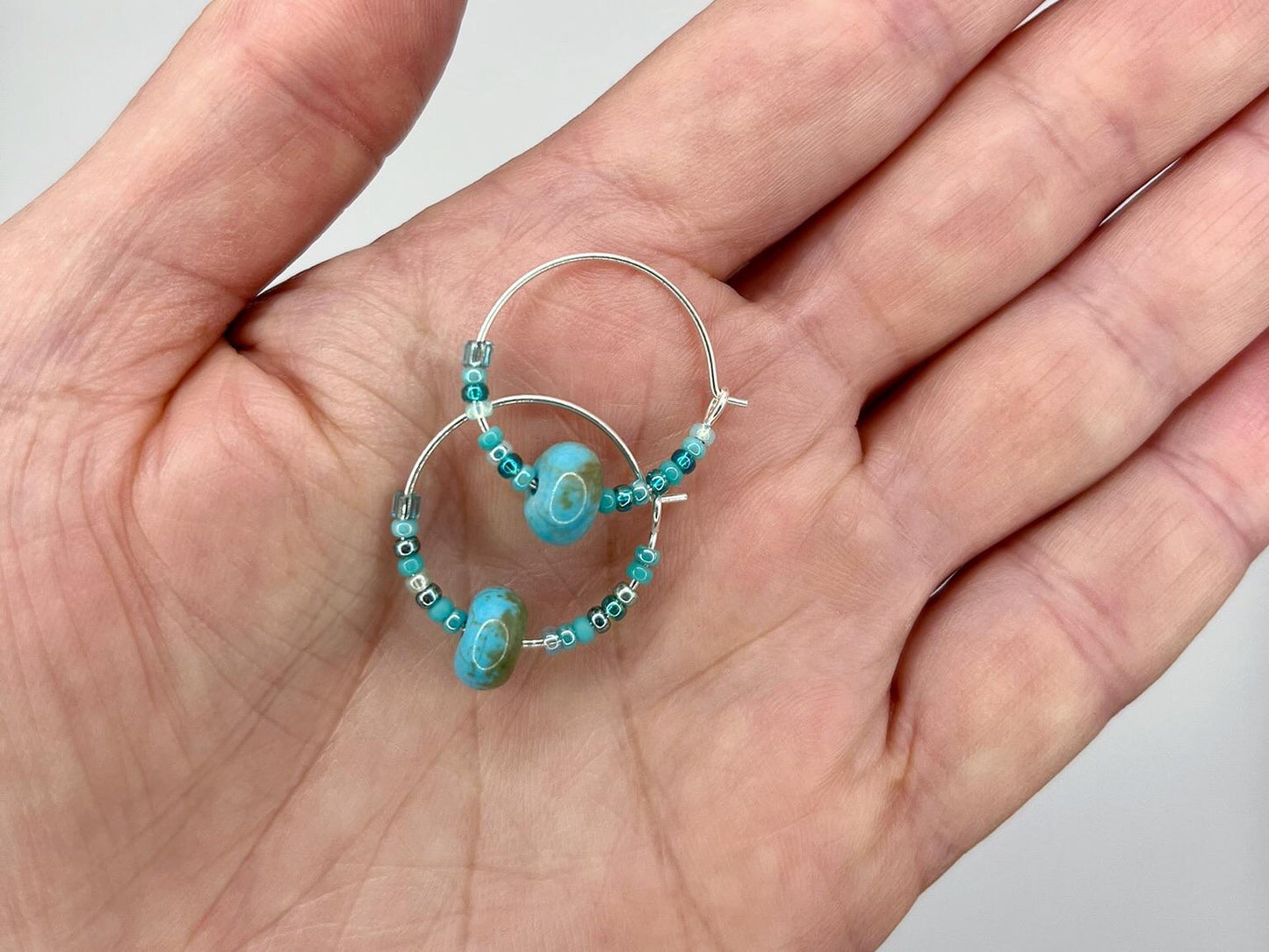 Hand holding a pair of turquoise beaded hoop earrings against a neutral background