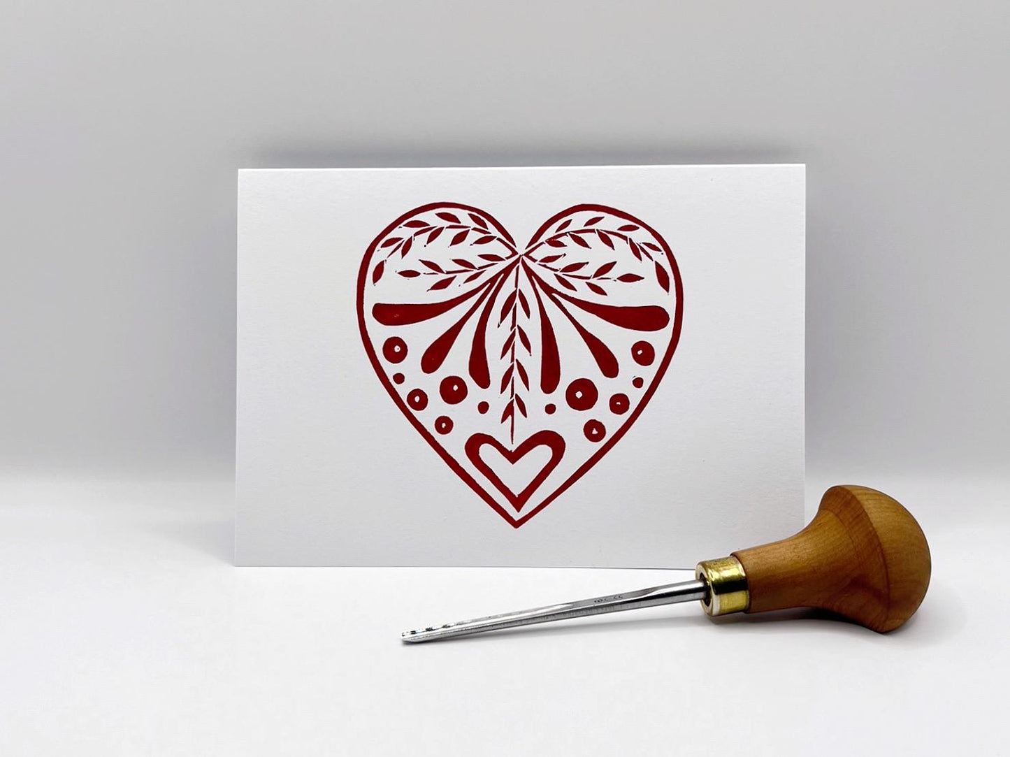 White card with an original lino printed red heart design stood on a white surface. Photo includes lino cutting tool.