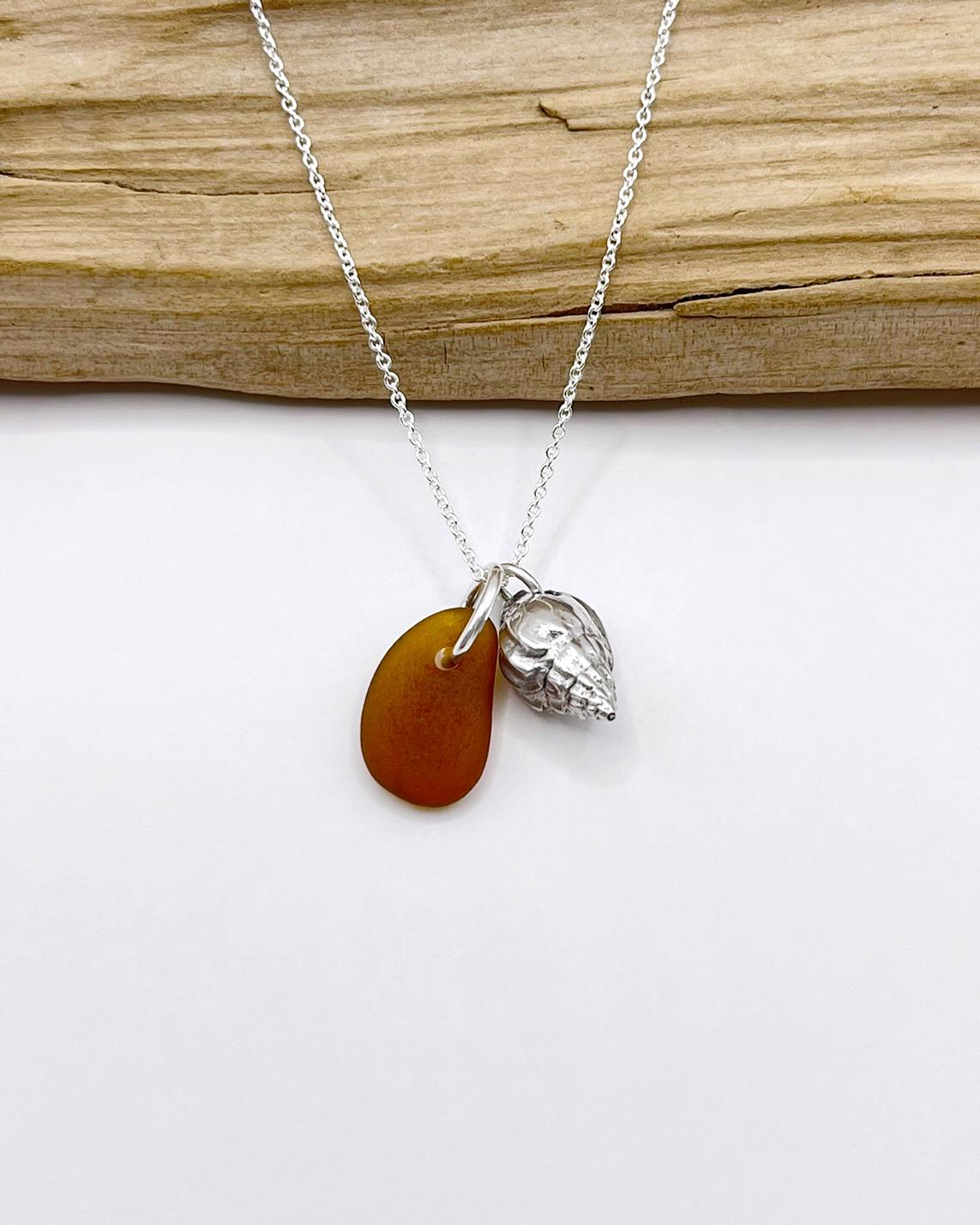 Necklace with burnt orange sea glass and silver shell pendant laid in front of driftwood on a white background