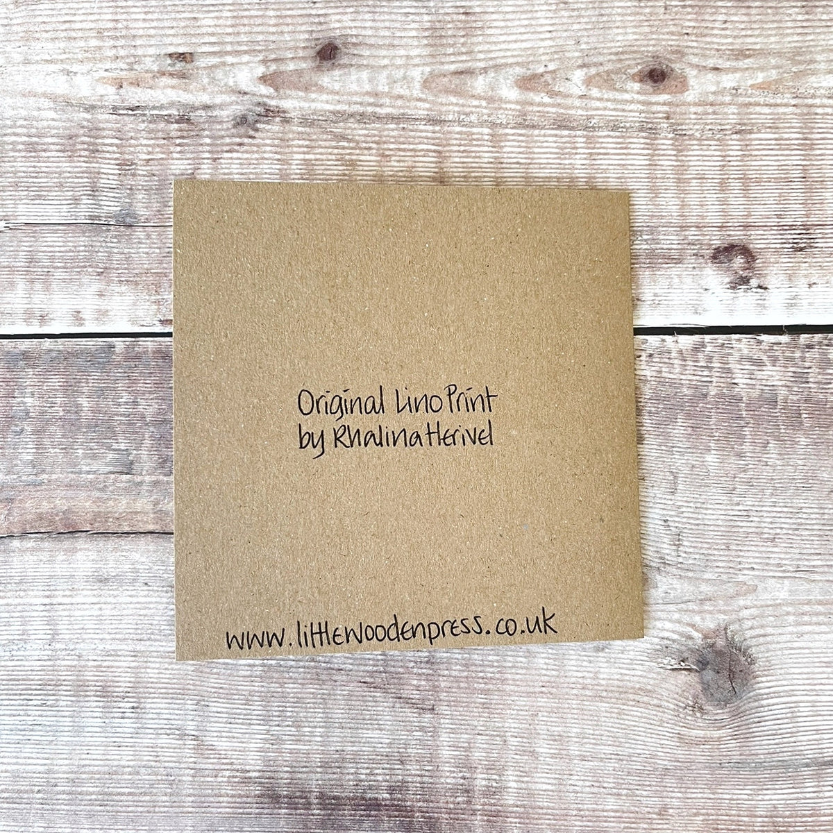 Showing the back of a square brown kraft paper card with black handwritten text reading 'Original Lino Print by Rhalina Herivel' and the website 'www.littlewoodenpress.co.uk' Laid on a wooden background.