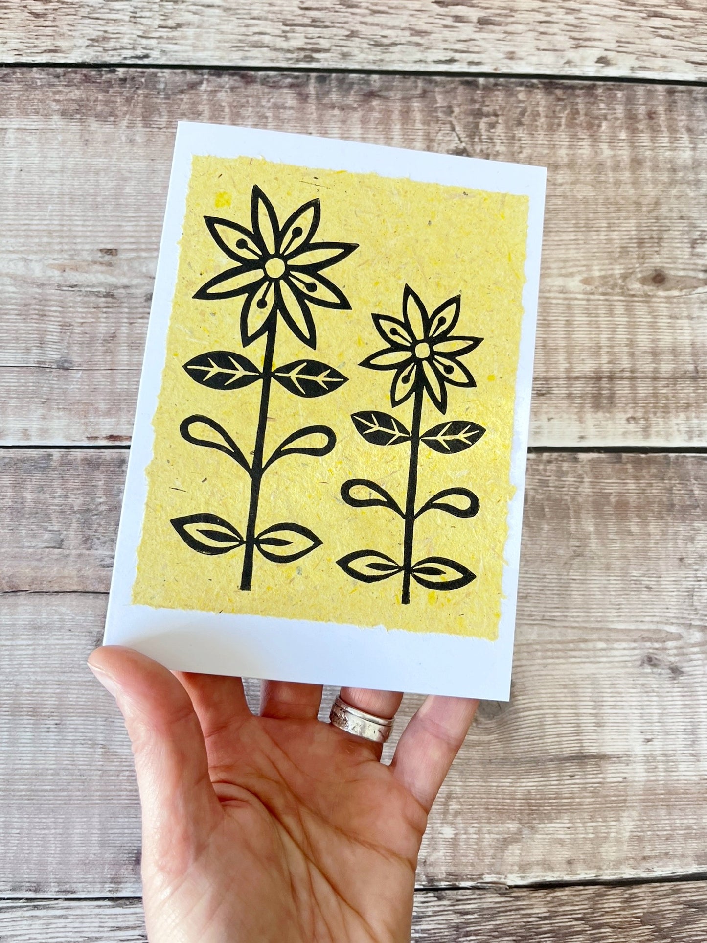 Lino print of two daisies in black ink on yellow handmade paper mounted on a white greetings card. Held in a hand against a wooden surface.