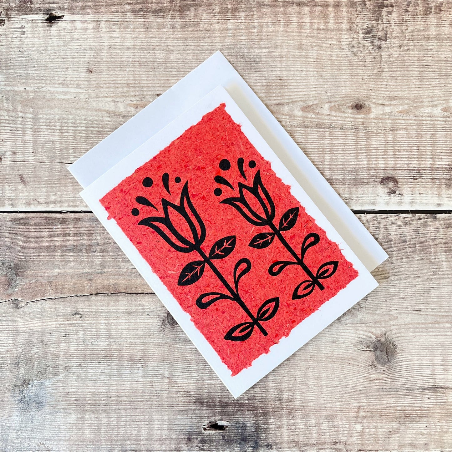 Lino print of two tulips in black ink on brick red handmade paper mounted on a white greetings card with matching envelope. Laid on a wooden surface.