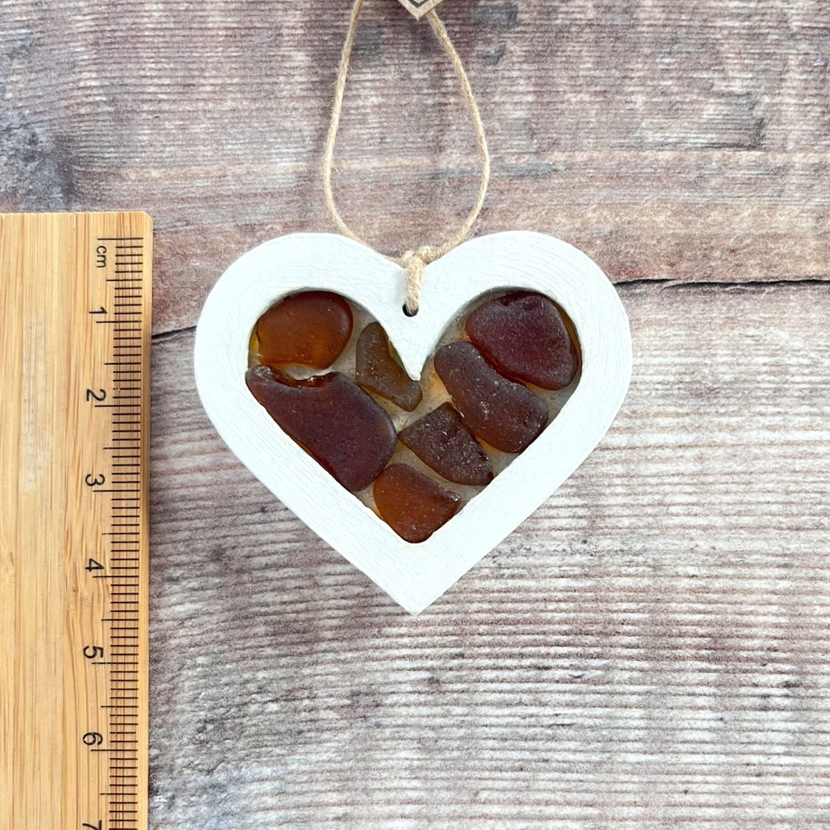 Heart-shaped sea glass hanging decoration next to a ruler for scale and laid on a wooden surface