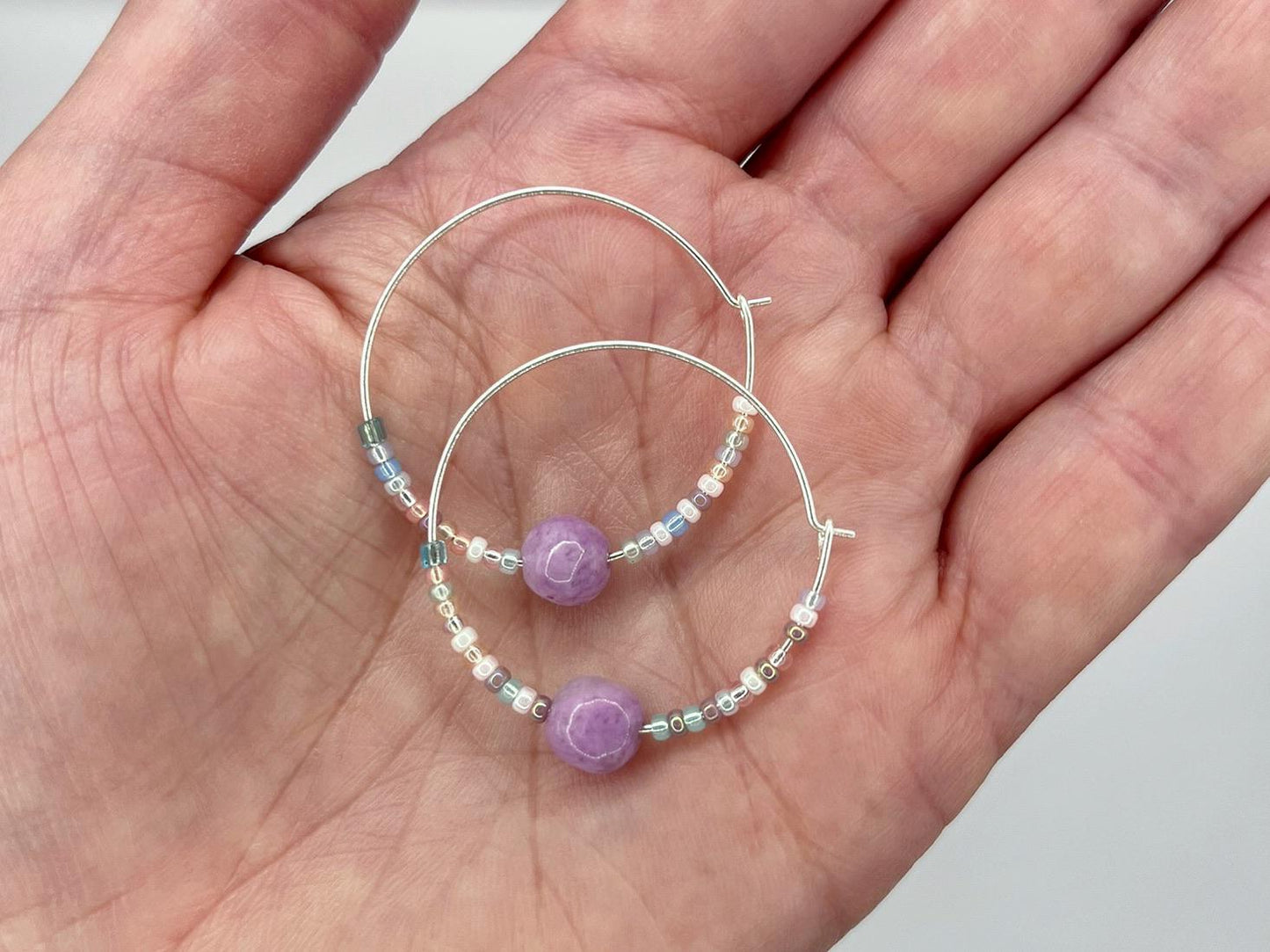 Pair of silver hoop earrings with lilac Jasper and seed beads held in a hand against a light surface.