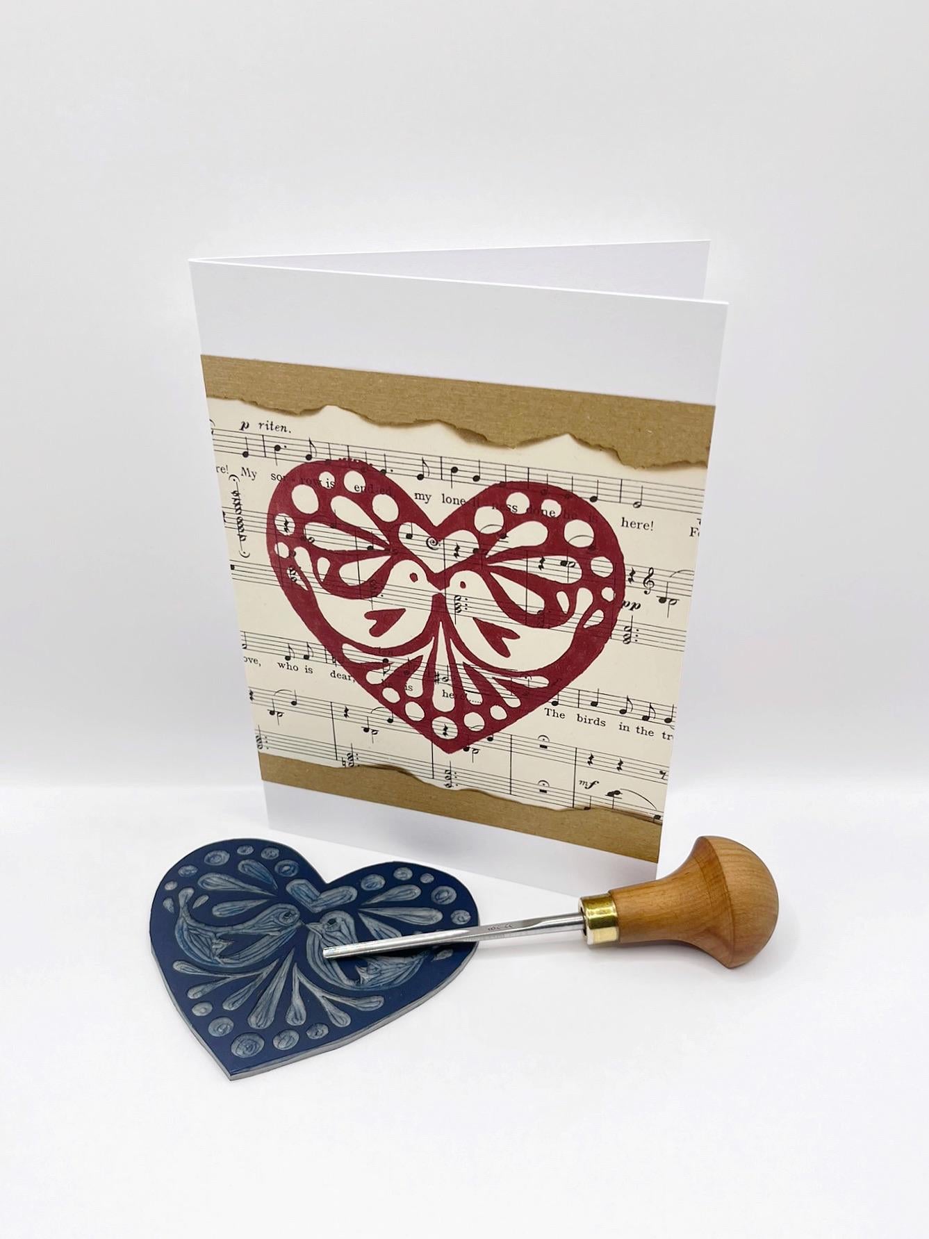 Card with red original lino printed heart design on sheet music. Photo included lino and cutting tool used stood against a white background.