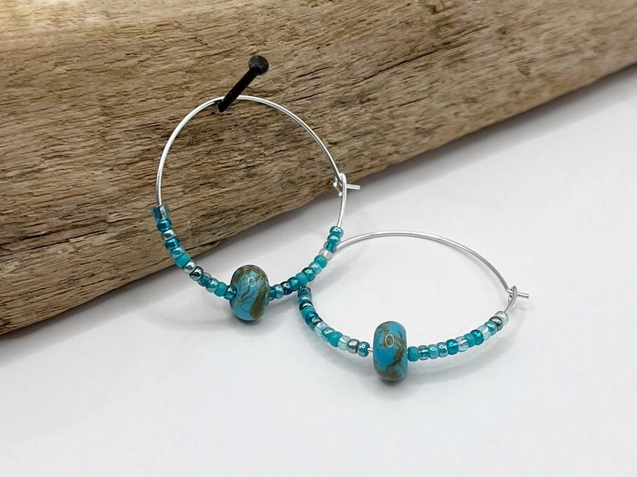 Pair of silver hoop earrings with turquoise stone and seed beads hanging on driftwood against a white surface