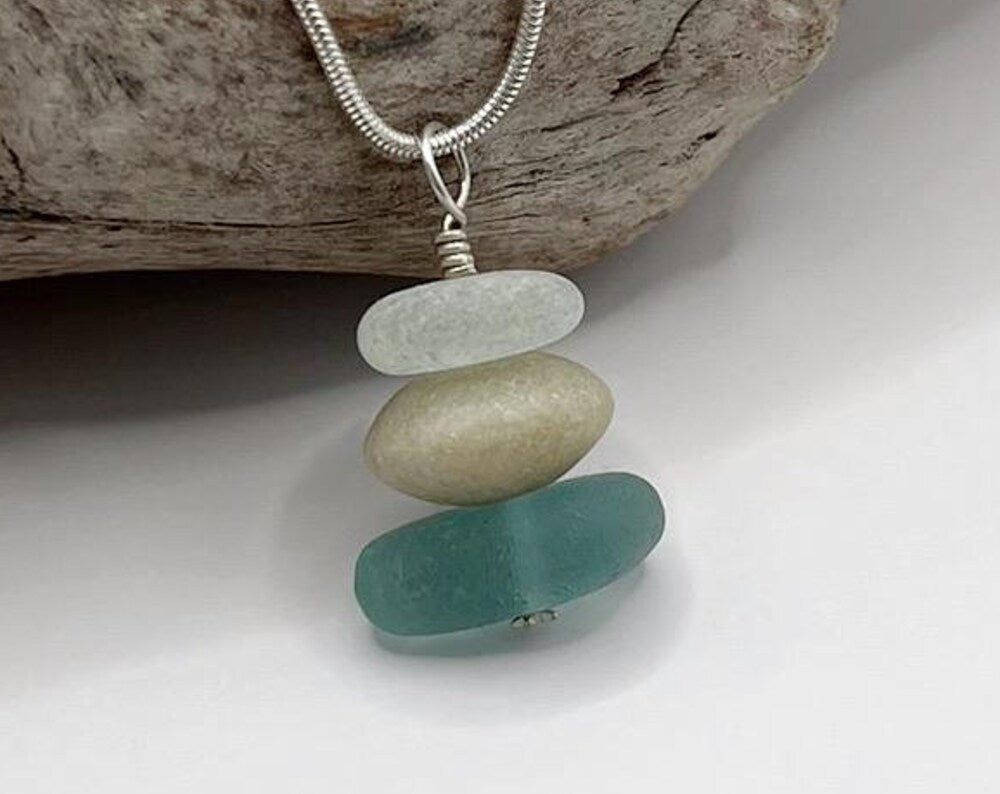 Close up of Cornish sea glass and Jade bead pendant on a silver snake chain necklace, leant against driftwood on a white surface.