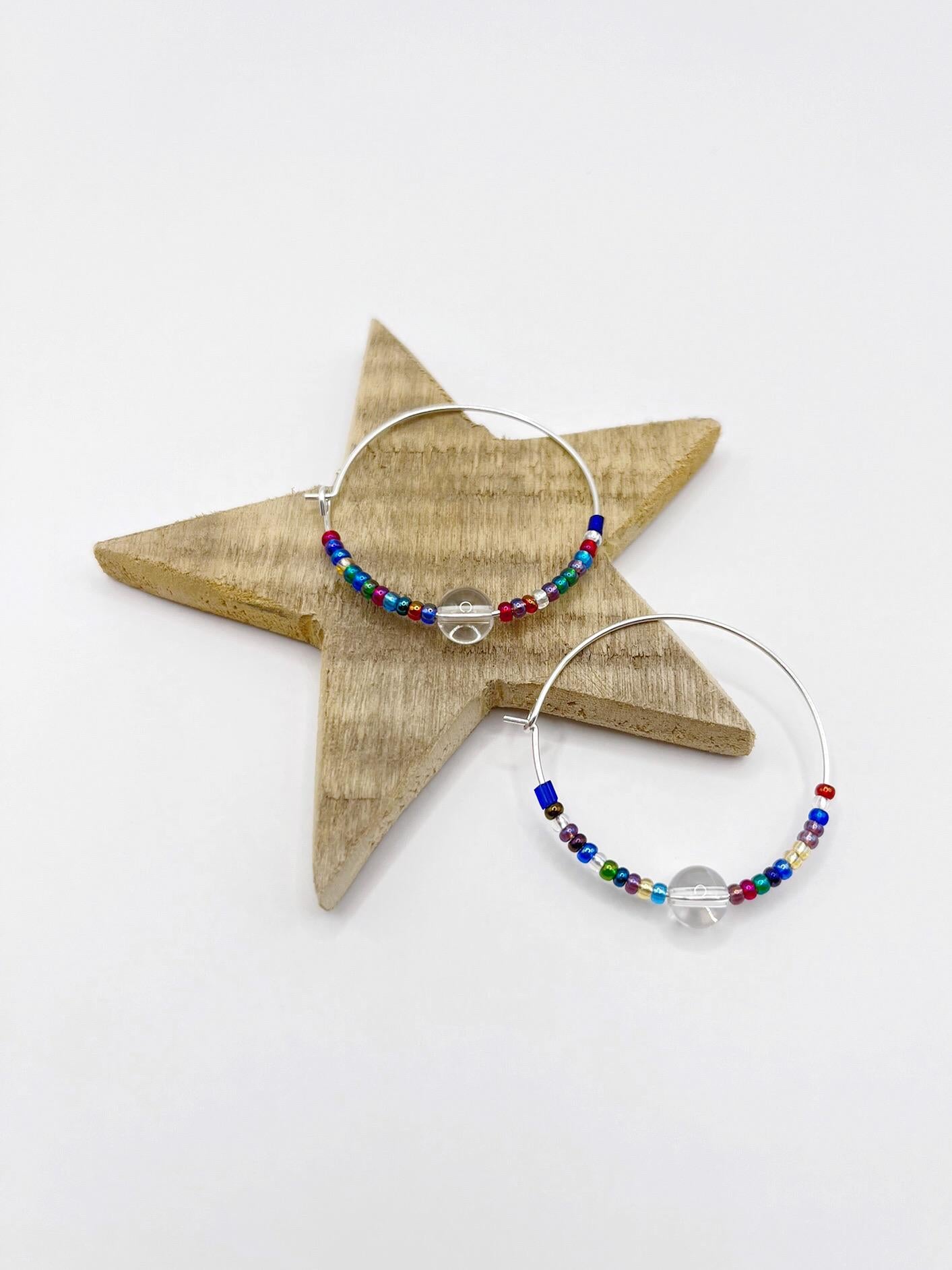 Rainbow seed bead and crystal hoop earrings laid on a wooden star against a white background.
