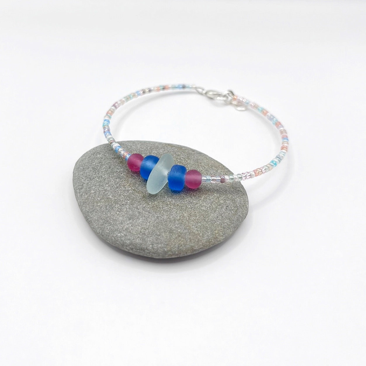 A pastel beaded bracelet with pink and blue frosted glass beads and a piece of sea foam coloured Cornish sea glass, finished with a silver plated lobster clasp and laid on a grey pebble.