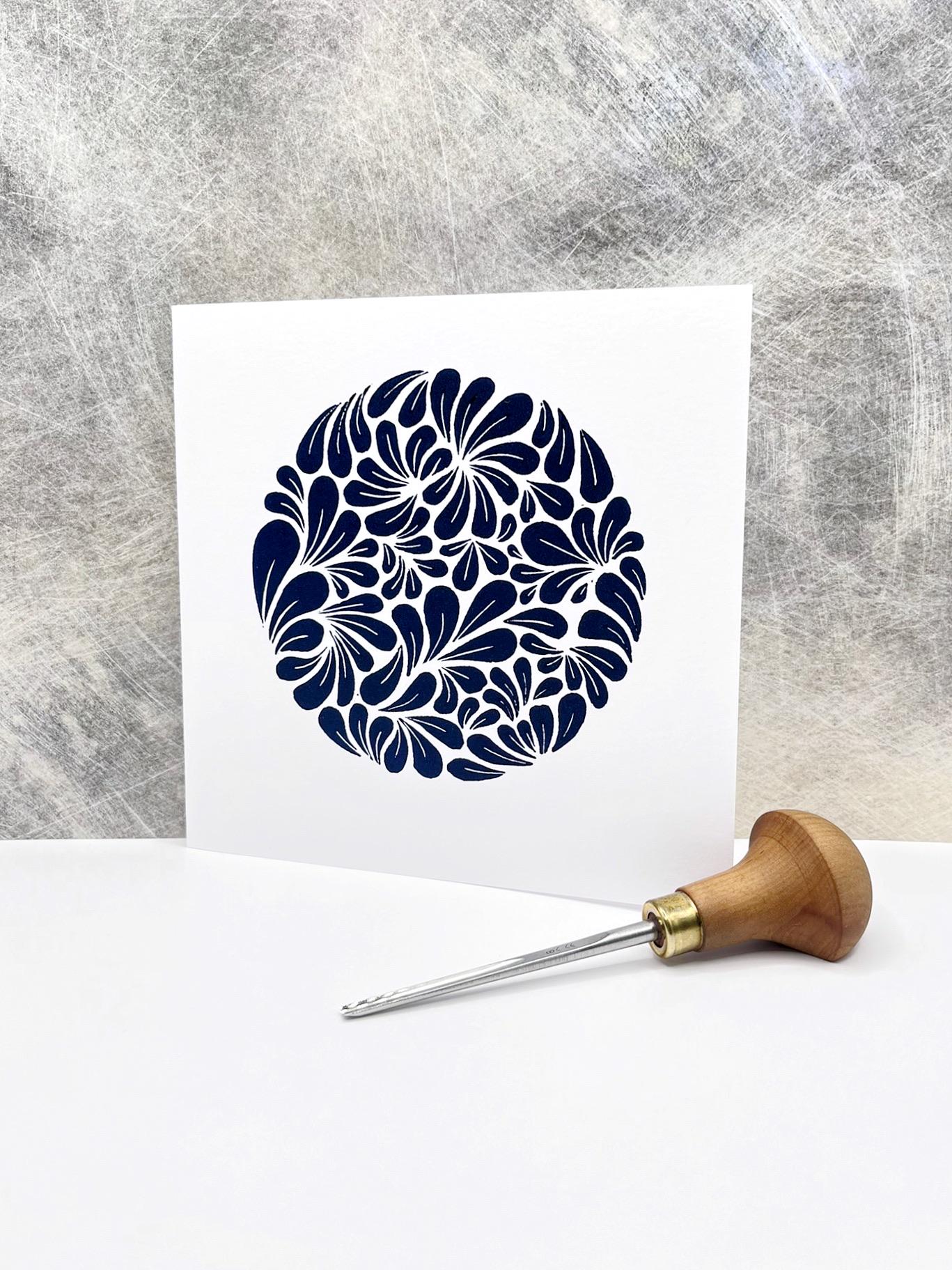 White card with a blue original lino print mussel shell pattern stood against  a textured grey background. Photo includes lino cutting tool