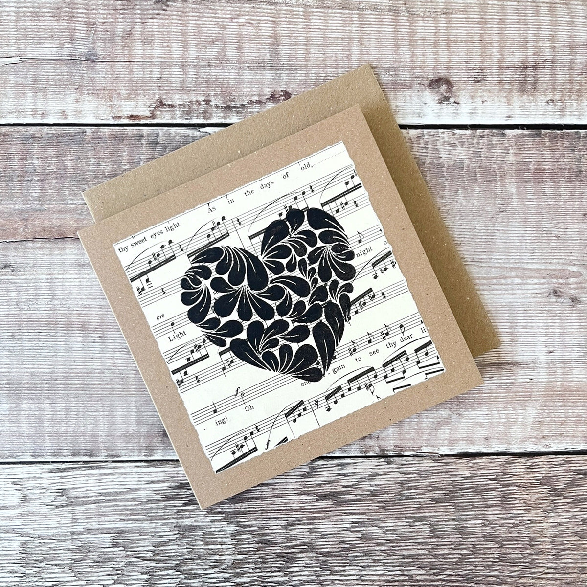 Square kraft greetings card with a lino printed heart design in black on music sheet paper mounted on the card. Laid on a wooden background.