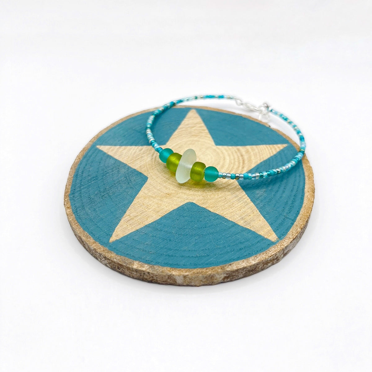 A turquoise beaded bracelet with lime and sea green frosted glass beads and a piece of sea foam coloured Cornish sea glass, finished with a silver plated lobster clasp and laid on a slice of wood with a blue star design.