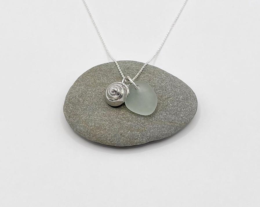 Necklace with a silver shell and seaglass pendant laid on a pebble against a white background