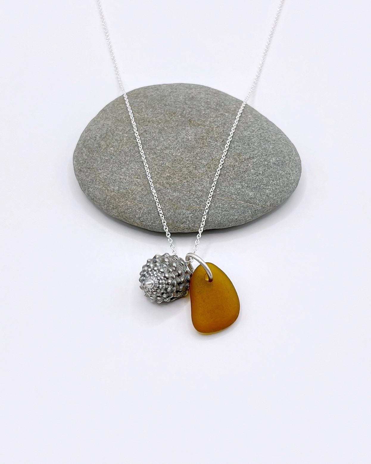 Necklace with burnt orange sea glass and silver shell pendant laid in front of a grey pebble against a white background