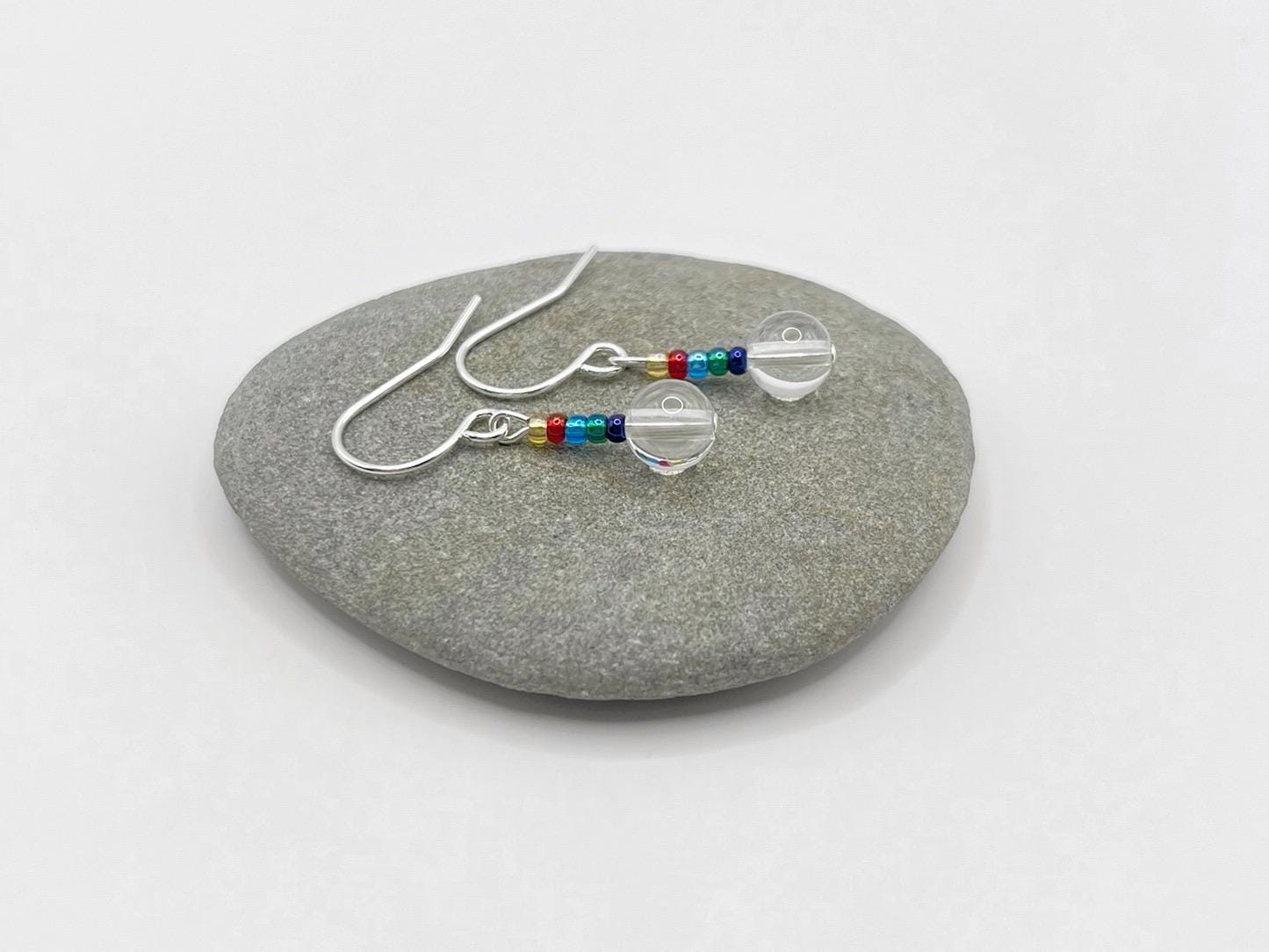 Silver earrings with clear crystal and rainbow seed beads laid on a grey pebble