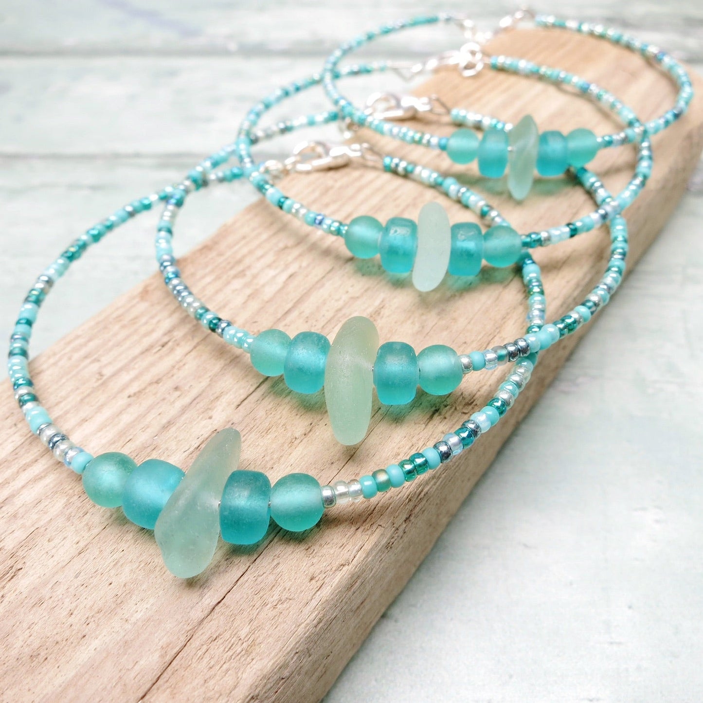 Four bracelets with pale sea foam sea glass and sea green and turquoise glass beads laid on driftwood against a pale blue background