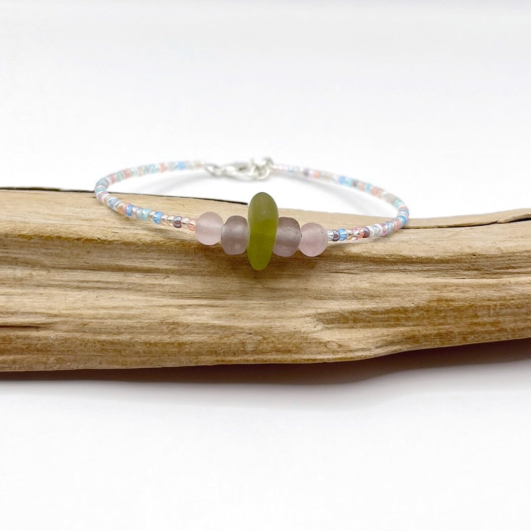 A pastel beaded bracelet with purple and lilac frosted glass beads and a piece of soft lime coloured Cornish sea glass, finished with a silver plated lobster clasp and laid on a piece of driftwood.