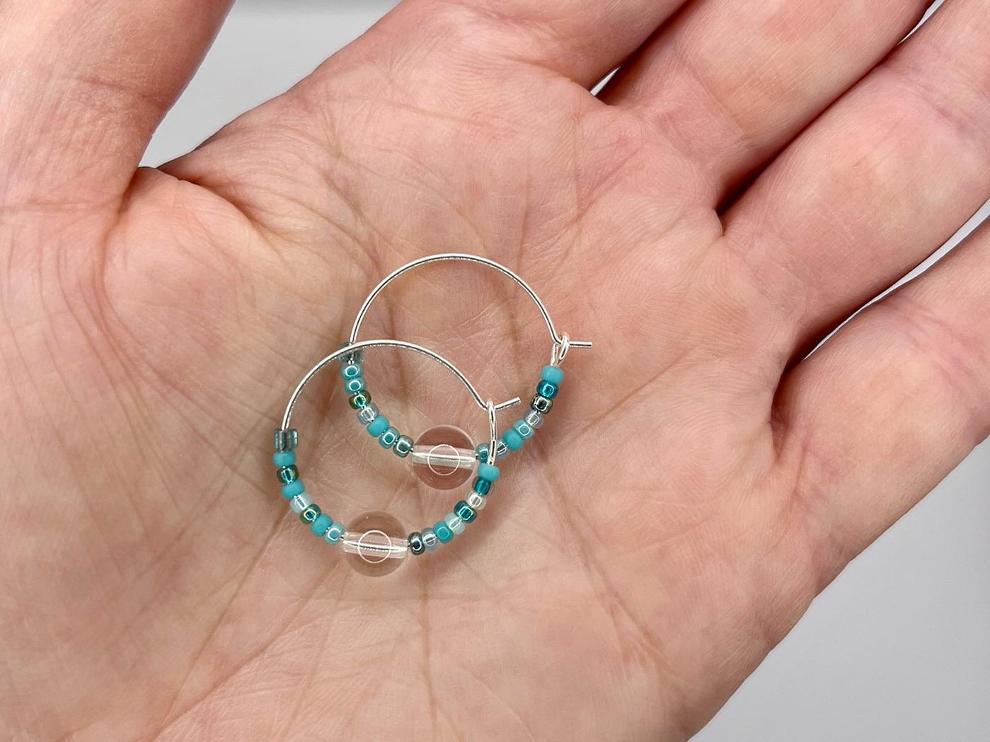 Silver hoop earrings with blue beads held in a hand against a neutral background
