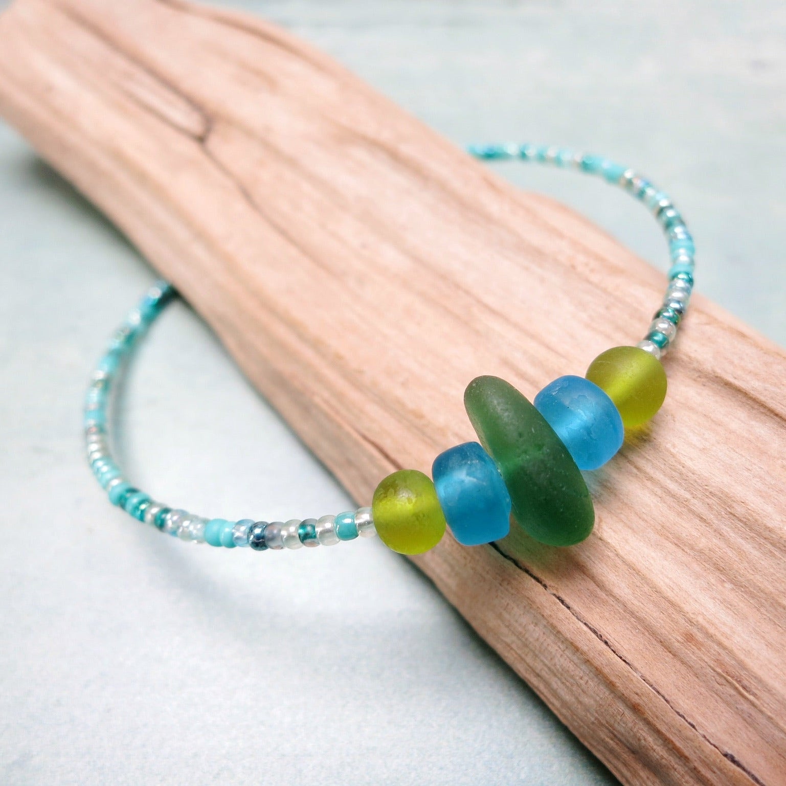 Close up of bracelet with green sea glass and turquoise and green glass beads laid on driftwood against a pale blue background