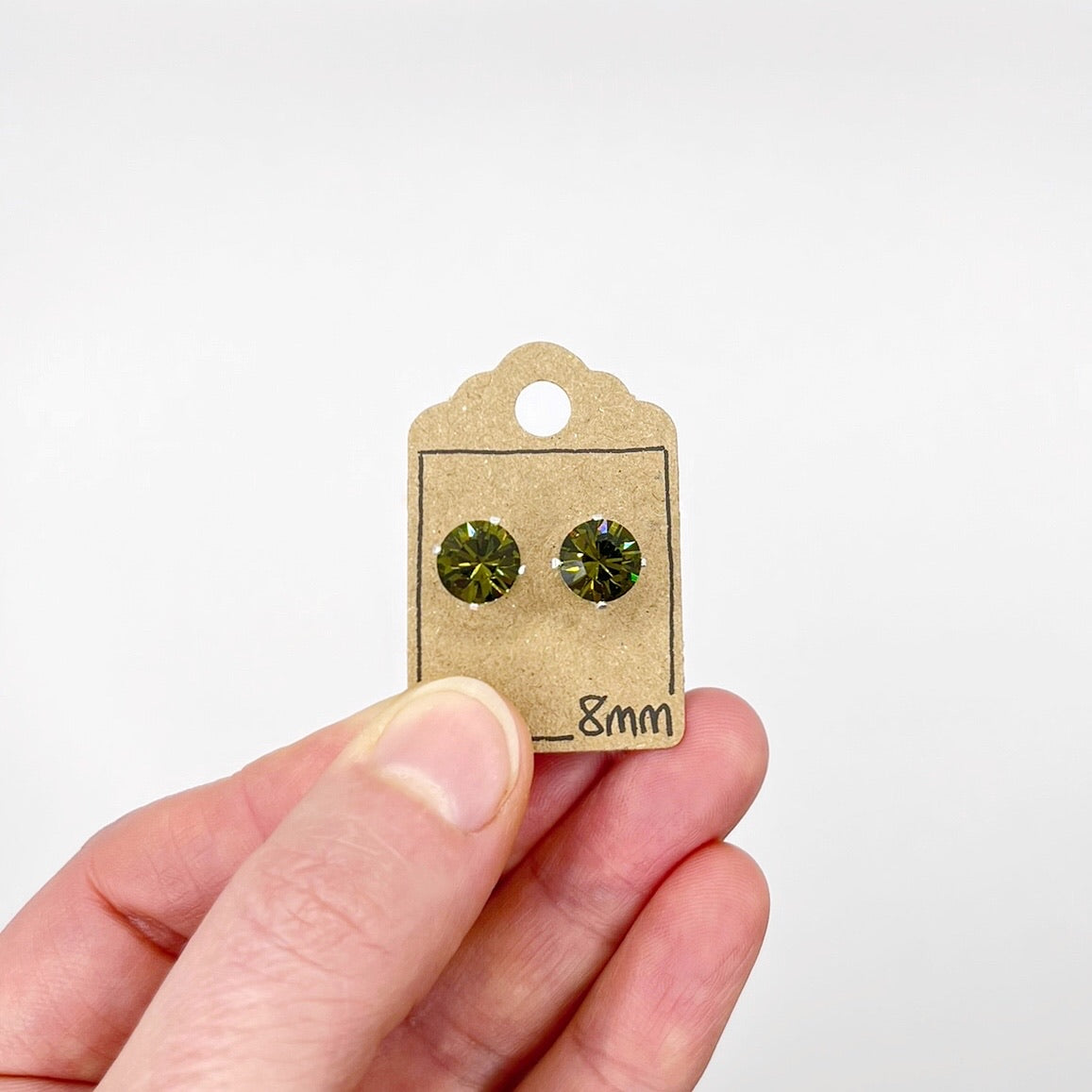 Hand holding a pair of olive green stud earrings on a cardboard backing with '8mm' text.
