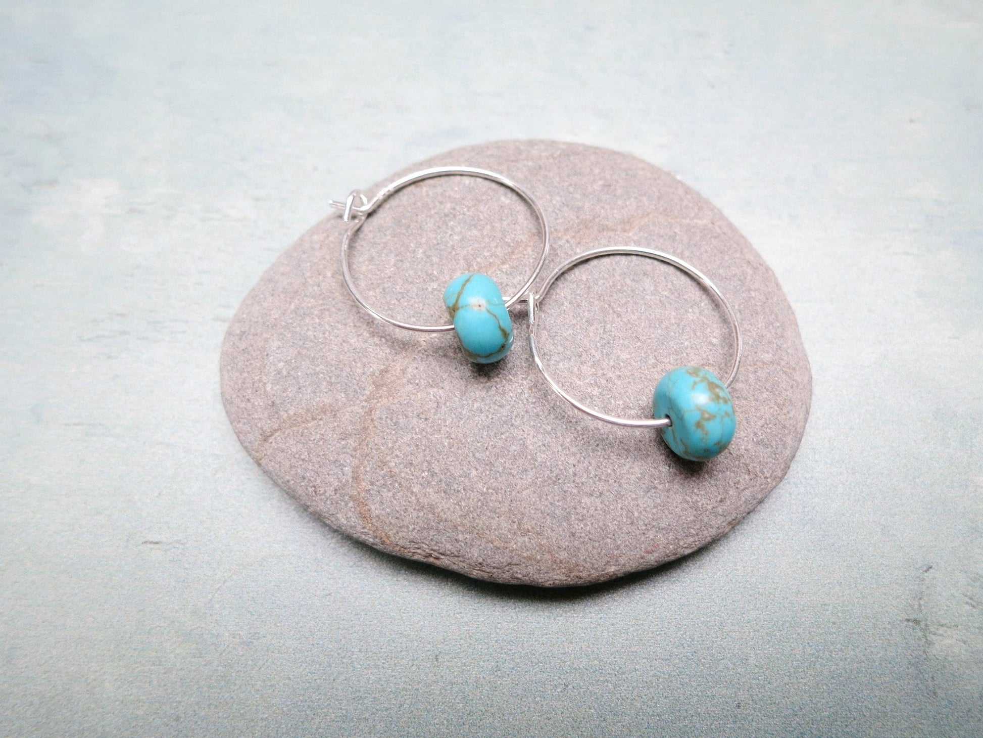 Silver hoop earrings with turquoise beads laid on a pebble against a pale green background