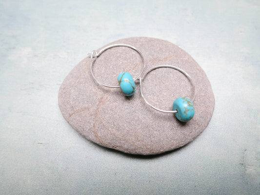 Silver hoop earrings with turquoise beads laid on a pebble against a pale green background