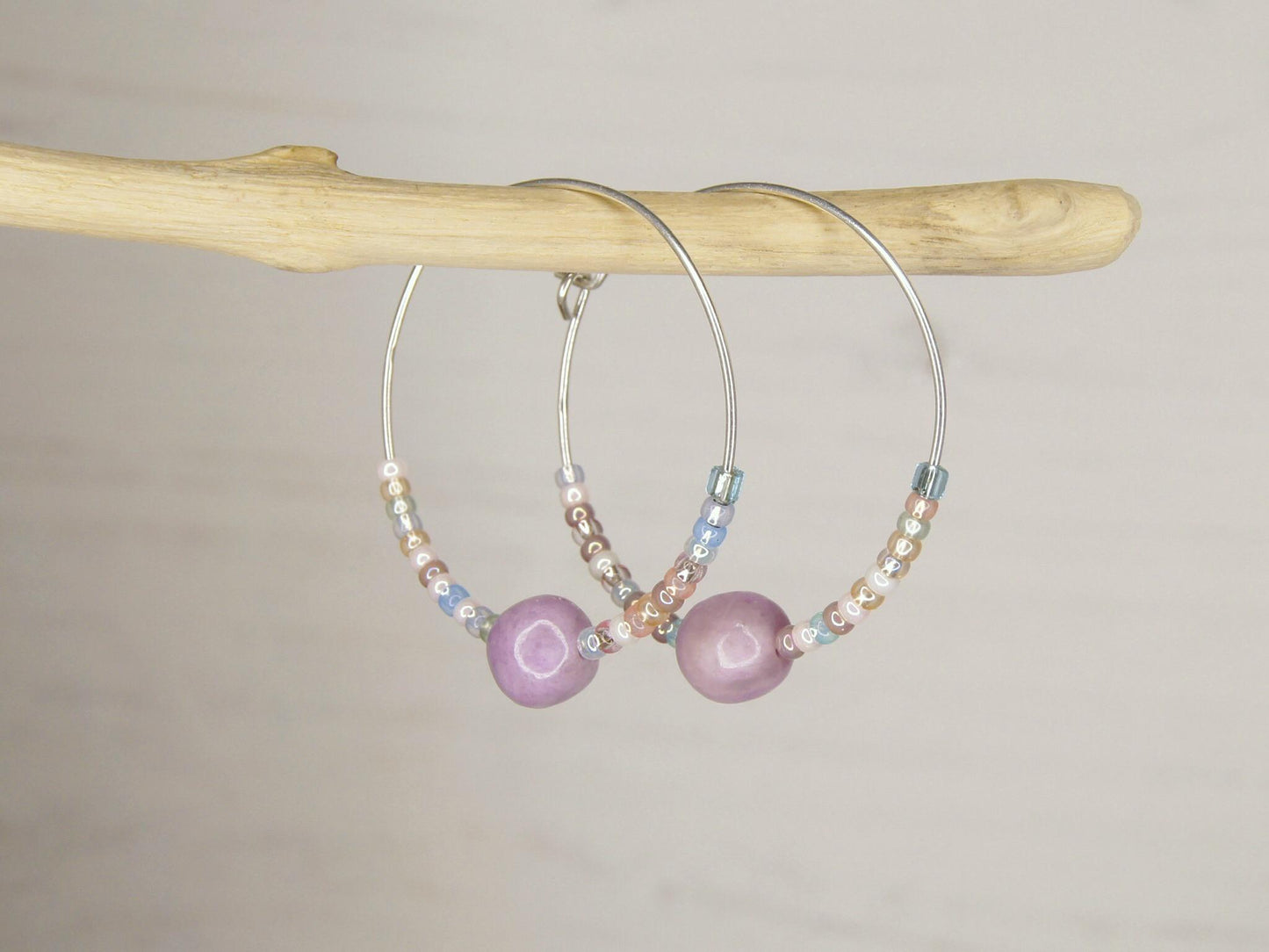 Pair of silver hoop earrings with lilac Jasper and seed beads hanging on driftwood against a light surface.
