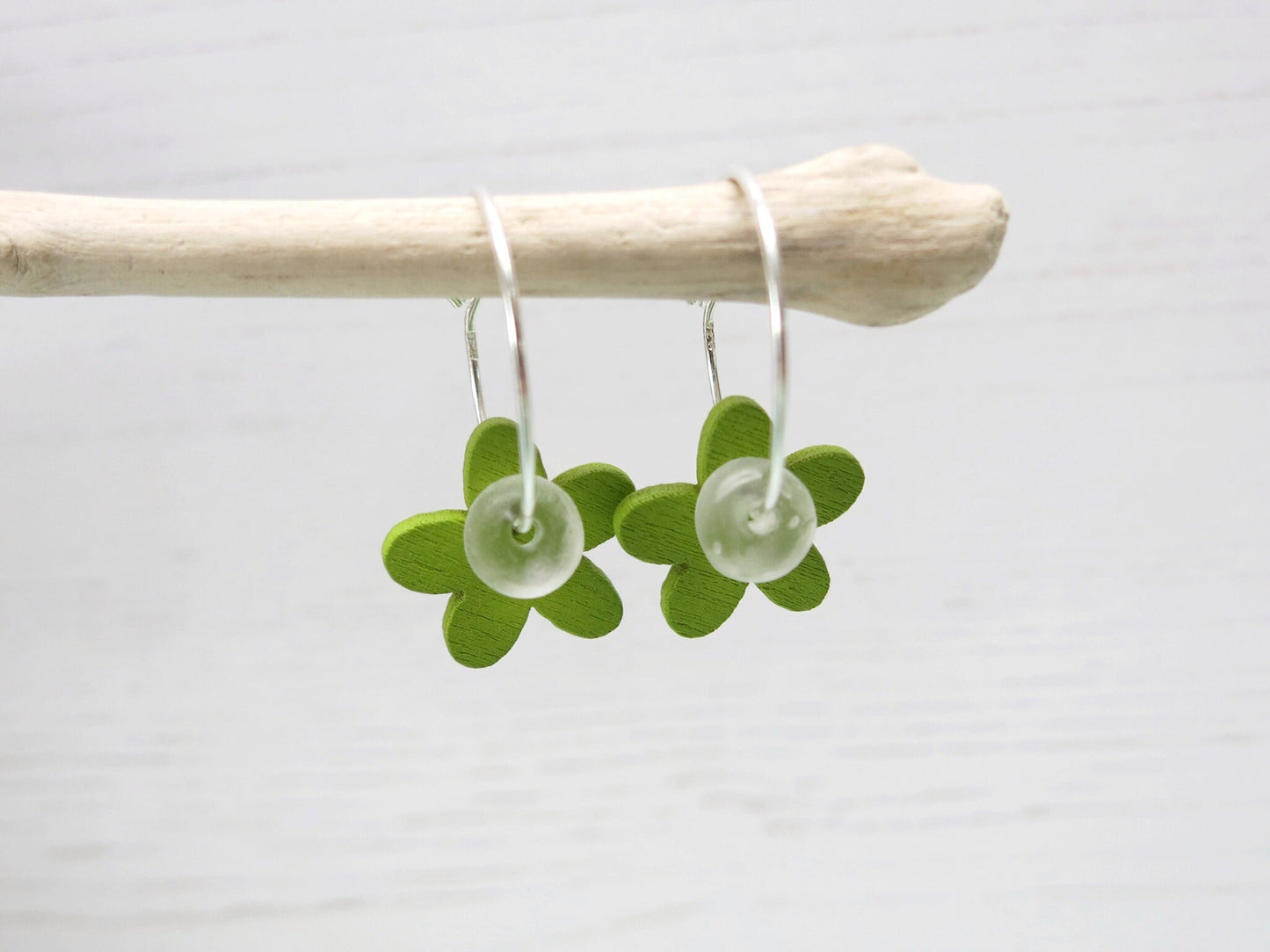 Silver hoop earrings with green wooden flowers and a glass bead hanging on driftwood against a white background
