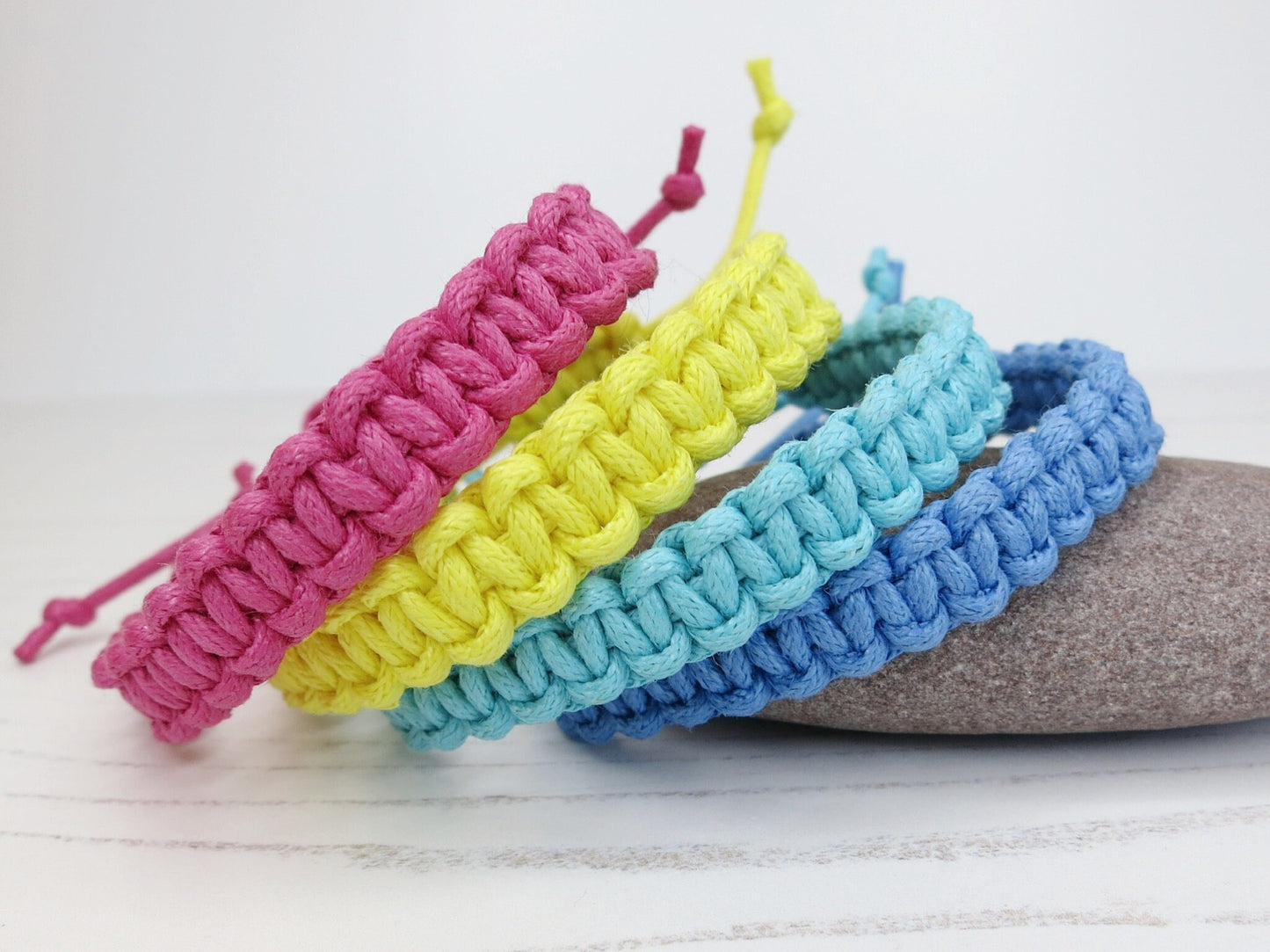 Four colorful macrame bracelets on a wooden surface with a pebble background.