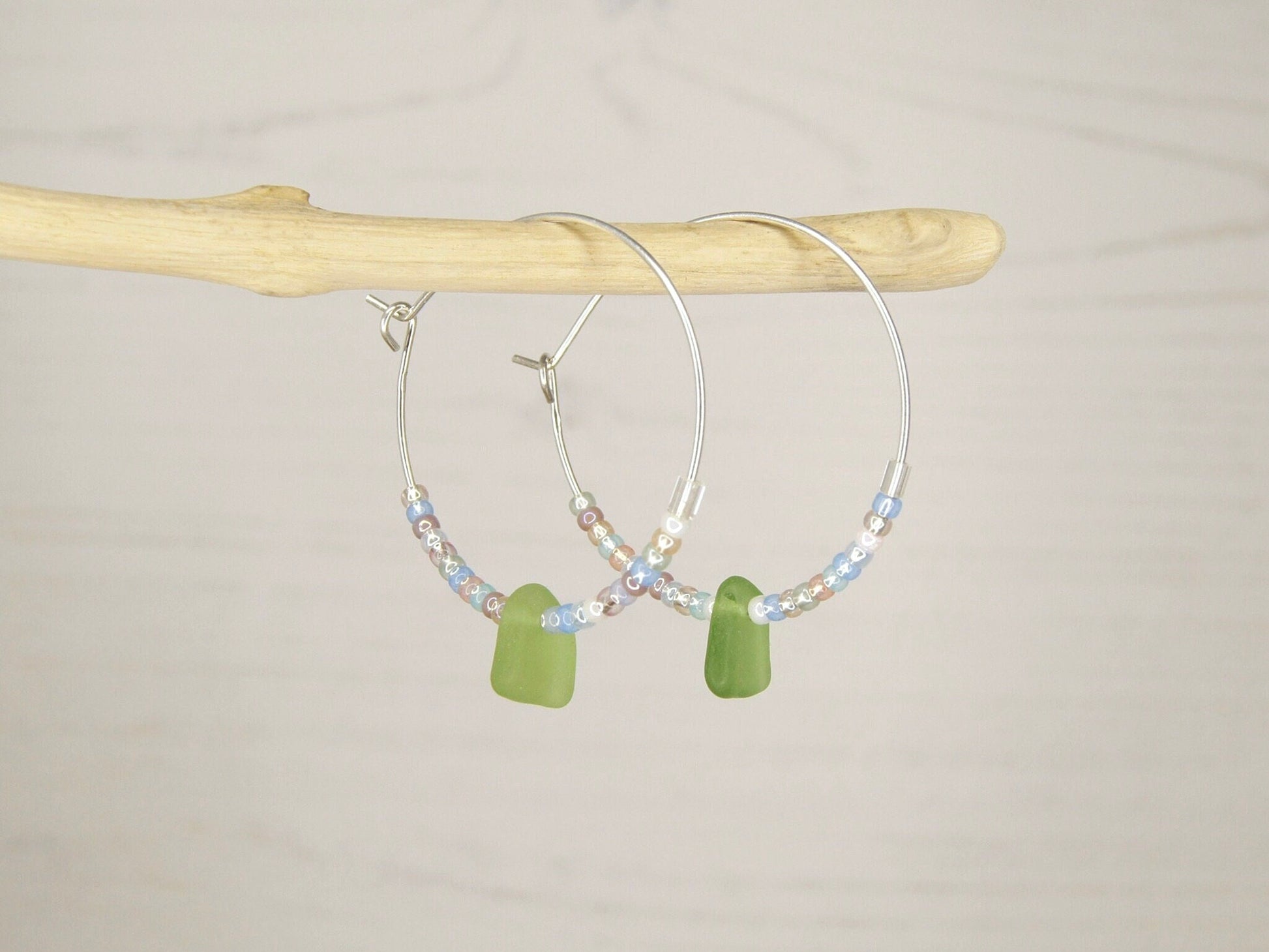 Hoop earrings with pastel seed beads and green sea glass hanging on driftwood against a pale background