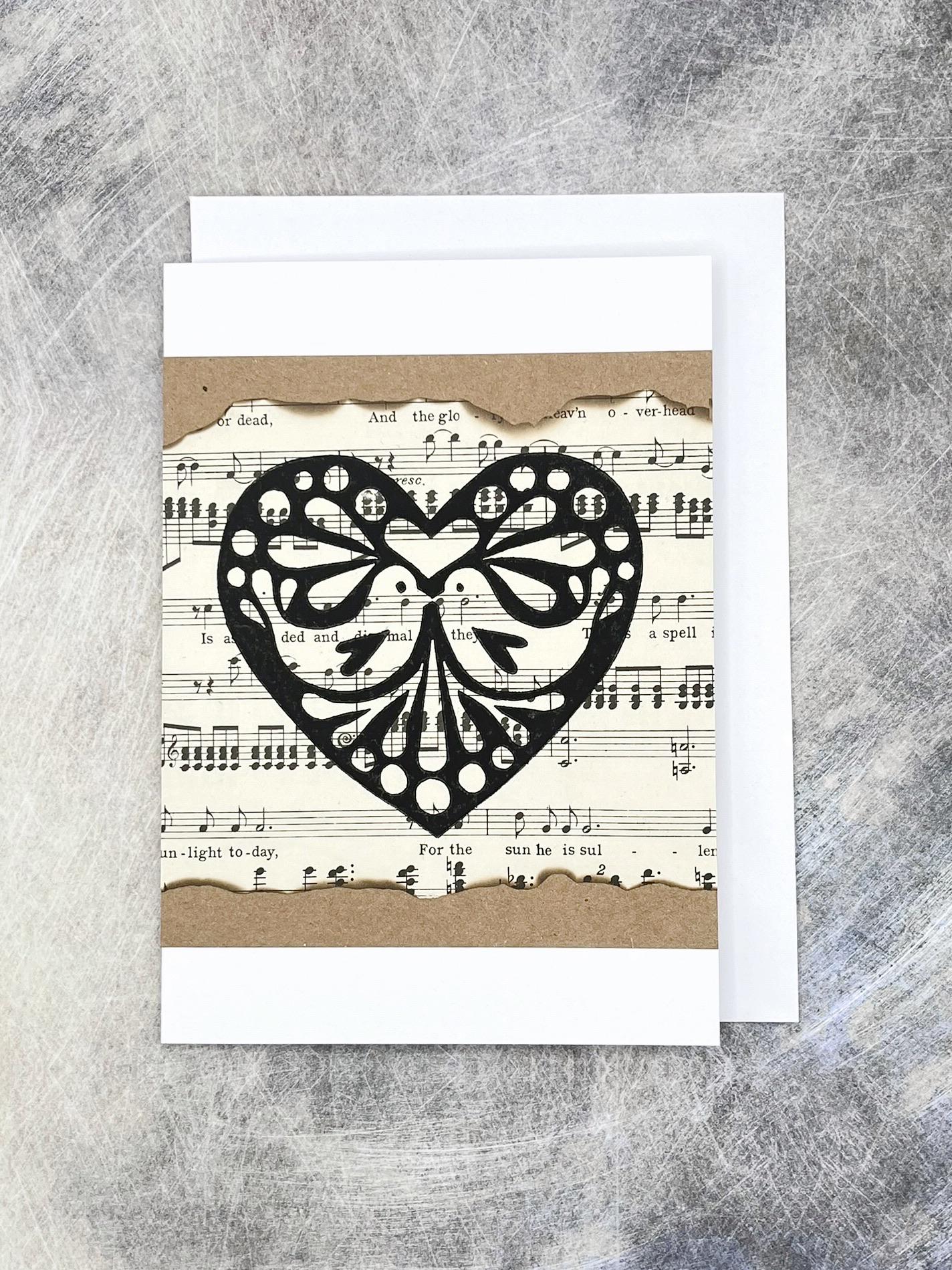 Greeting card with an original lino printed black heart design on sheet music, laid on a textured background.