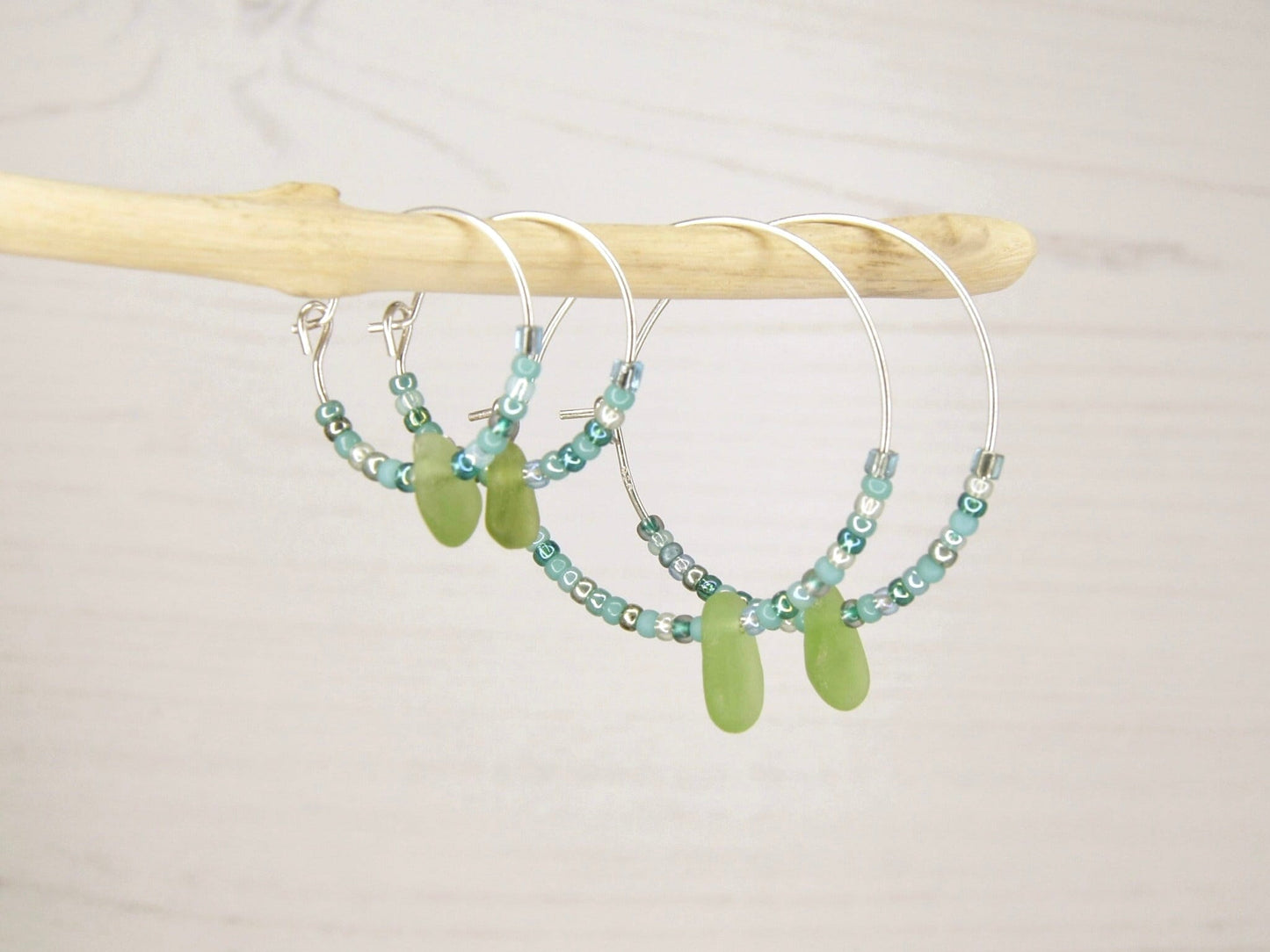 Hoop earrings with green sea glass and turquoise seed beads hanging on driftwood against a pale background