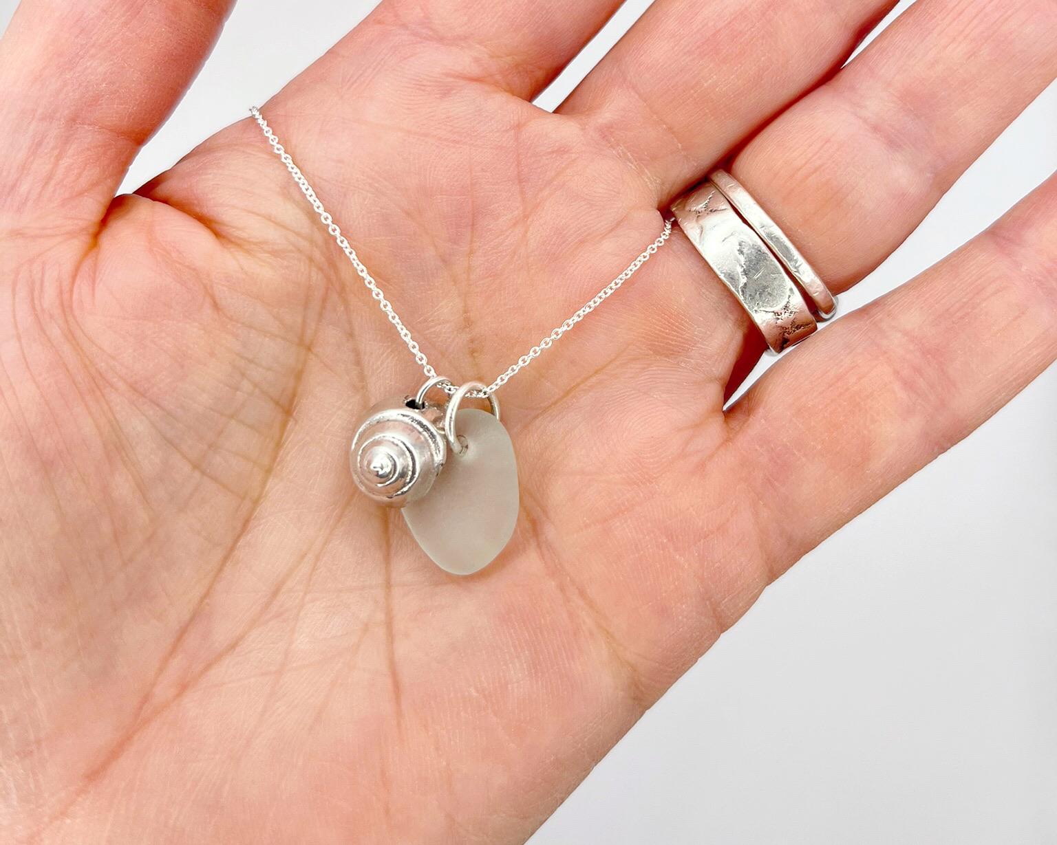 Hand holding a silver necklace silver shell and sea glass pendant against a neutral background