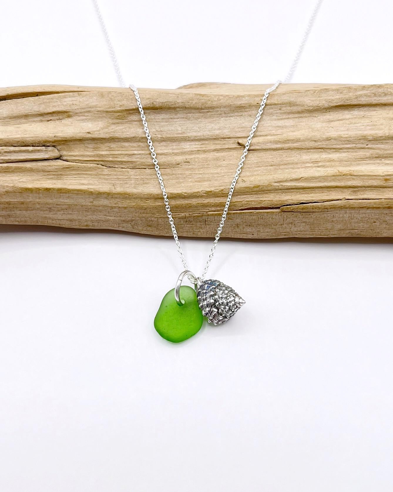 Necklace with green sea glass and silver shell pendant laid in front of driftwood on a white background