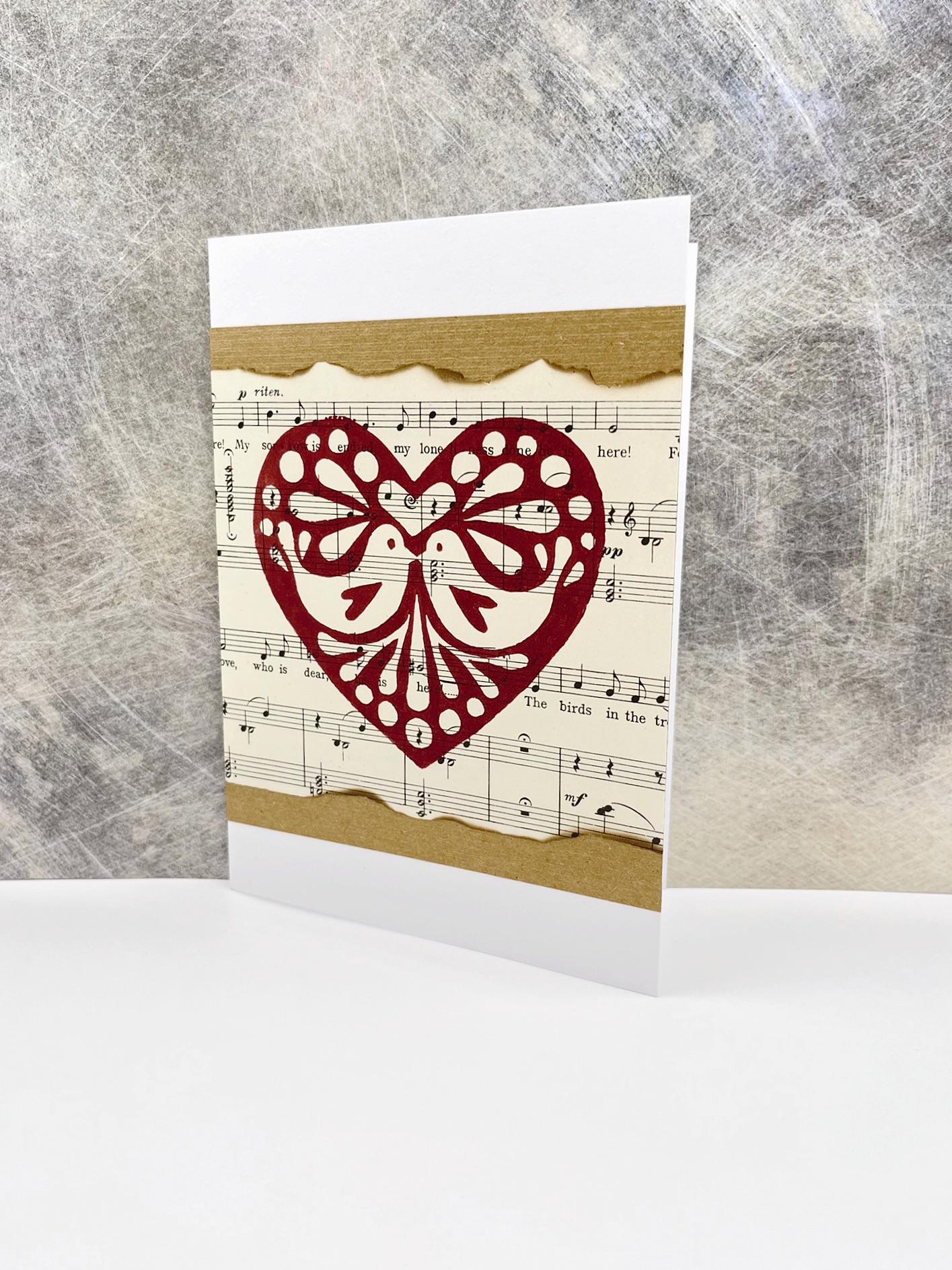 Greeting card with original lino printed heart design on sheet music stood against a textured background