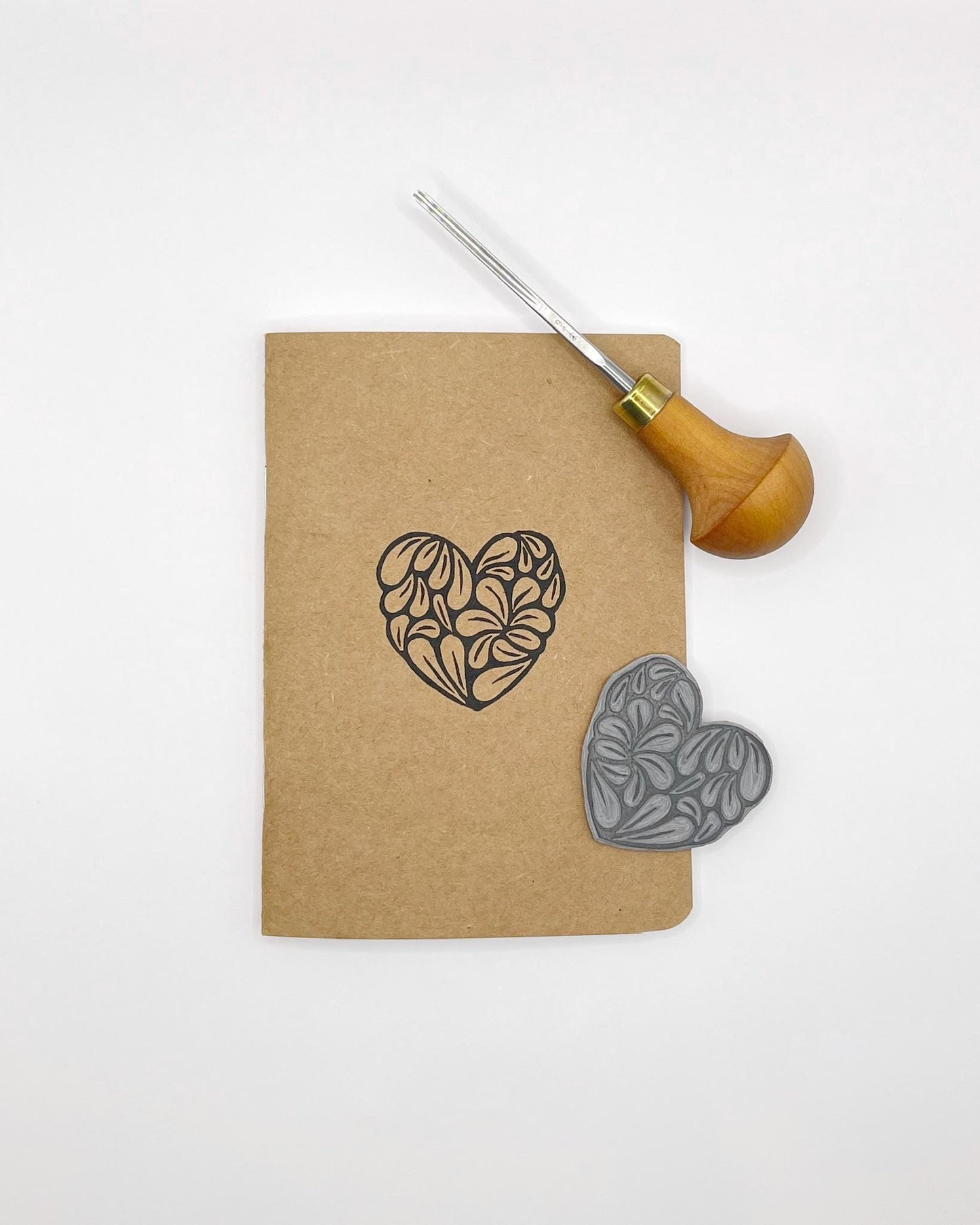 Kraft paper notebook with an original lino print heart, laid on a white background. Included in the photo is the lino and cutting tool used.