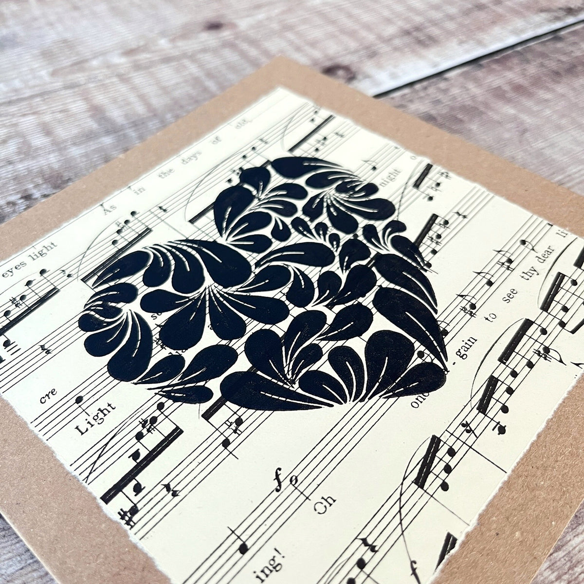 Close up of a square kraft greetings card with a lino printed heart design in black on music sheet paper mounted on the card. Laid on a wooden background.