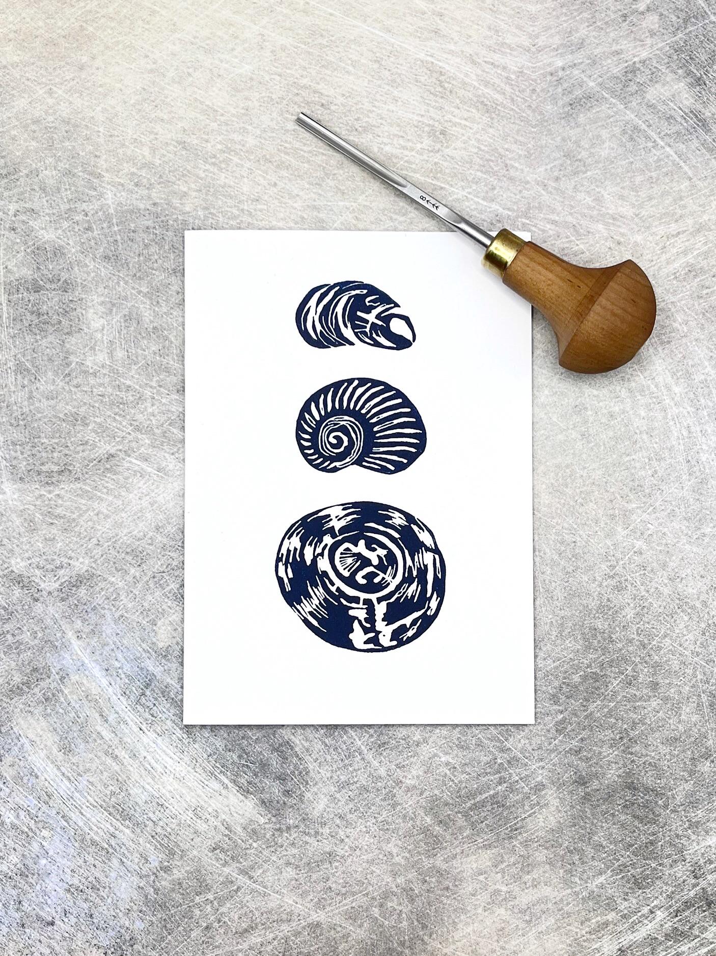 Three blue shells on a white card laid on a grey textured background. Included in the photo is the lino cutting tool used.