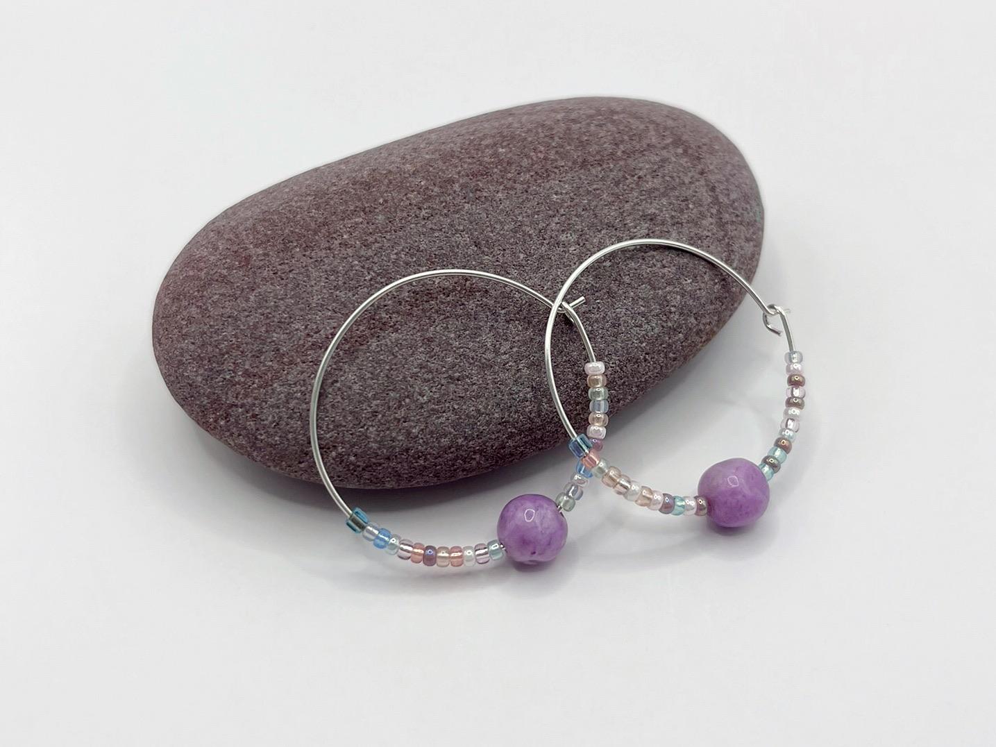 Pair of silver hoop earrings with lilac Jasper and seed beads laid against a purple pebble on a white background.