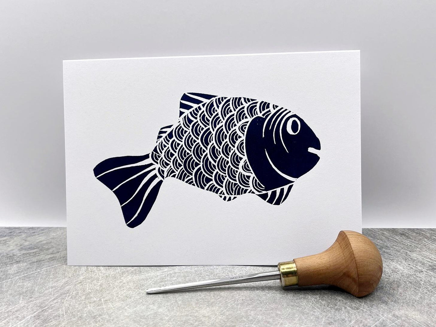 White card with an original lino printed blue fish, stood against a white background. Photo includes the lino cutting tool used.