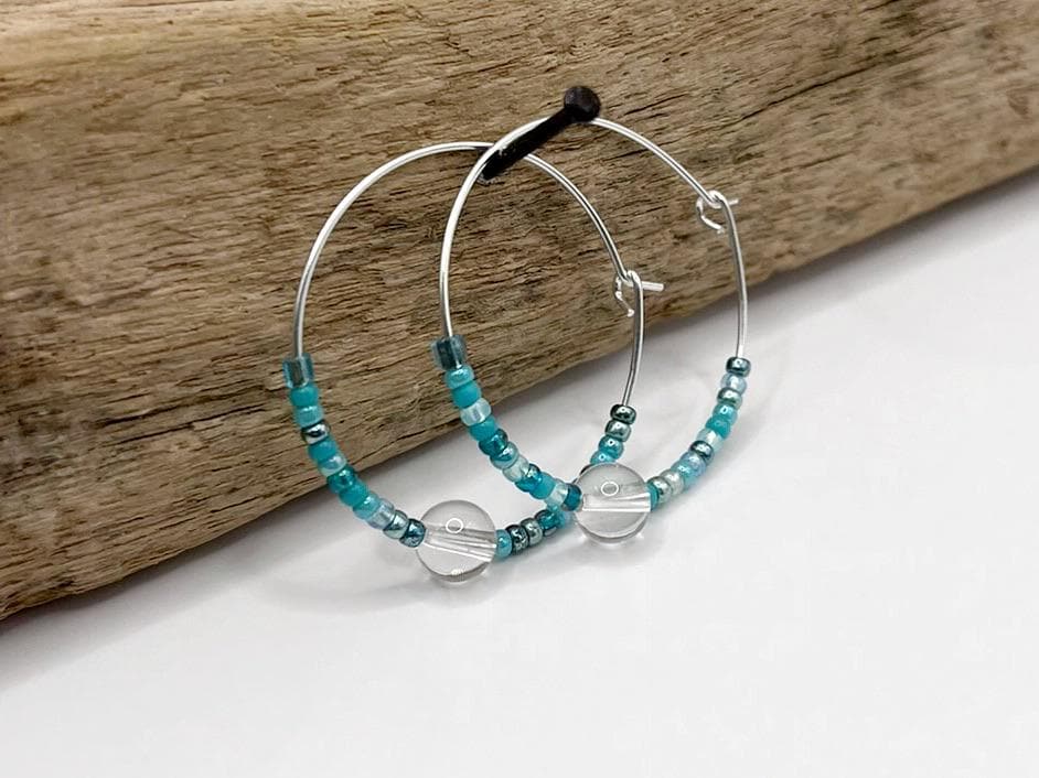 Silver hoop earrings with turquoise seed beads and crystal hanging on driftwood against a white background