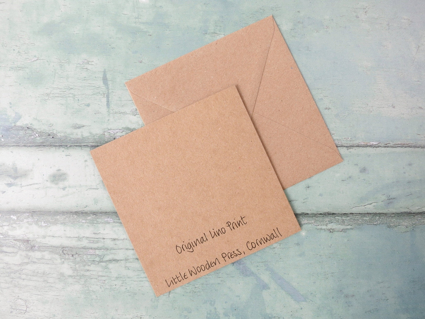 Showing the back of the Kraft card with the text 'Original Lino Print Little Wooden Press, Cornwall' and a matching envelope laid on a textured surface.