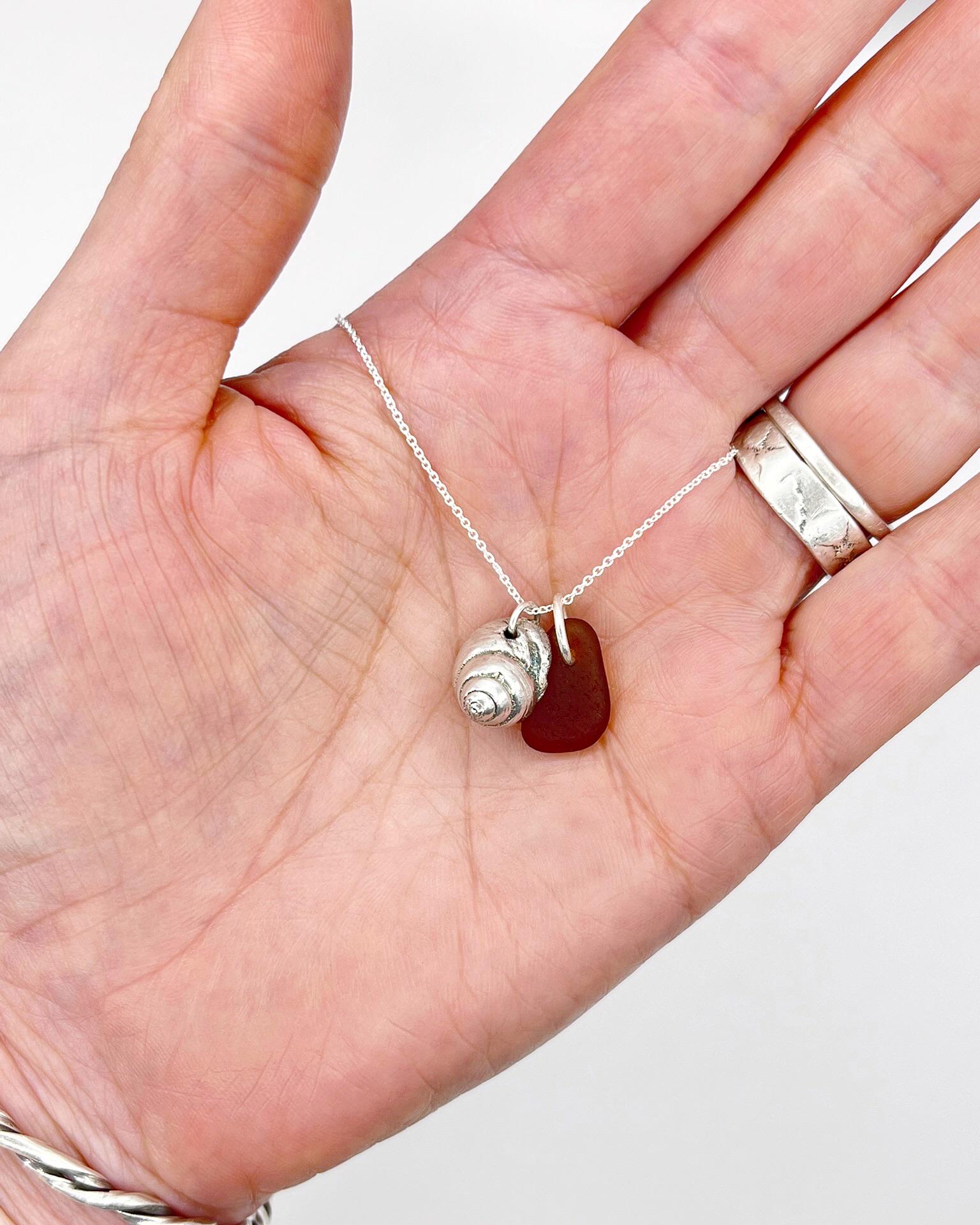 Hand holding a silver necklace with burnt orange sea glass and a silver shell pendant against a white background