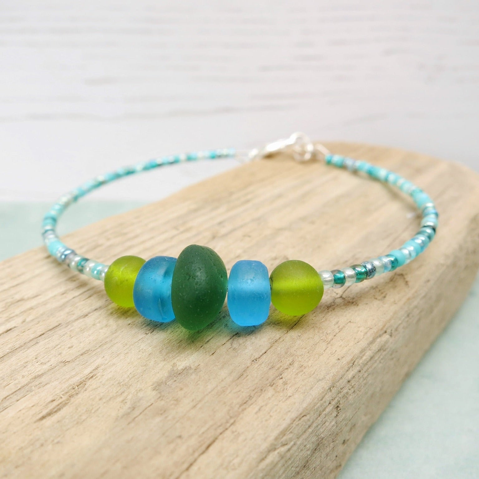 Close up of bracelet with green sea glass and turquoise and green glass beads laid on driftwood against a pale blue background
