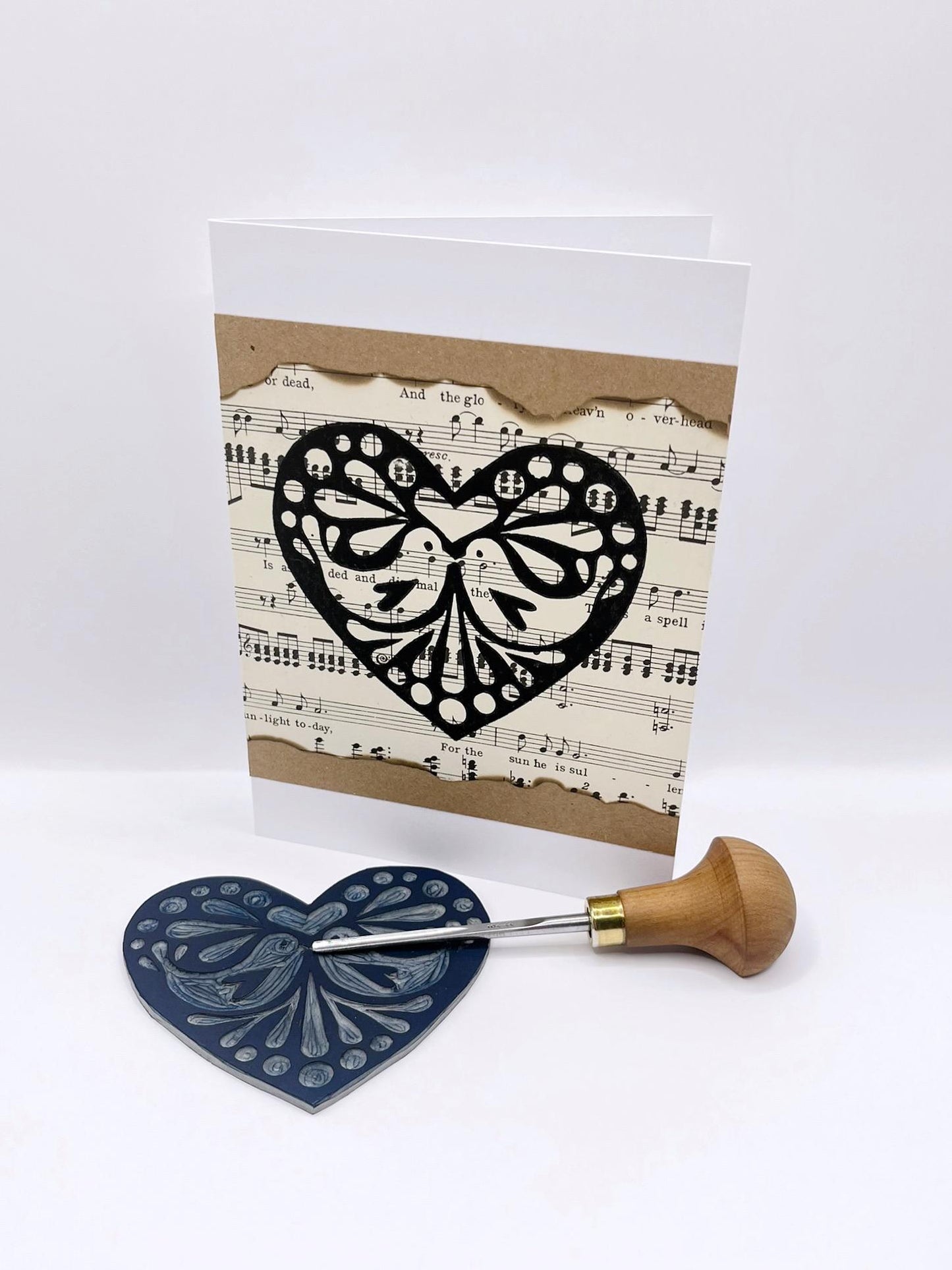 Card with black heart design on sheet music, stood against a white background. Photo includes lino and cutting tool.