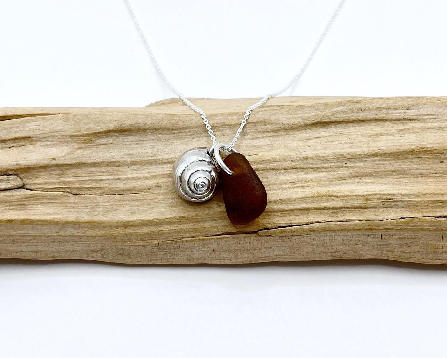 Silver necklace with burnt orange sea glass and a silver shell pendant laid on driftwood against a white background