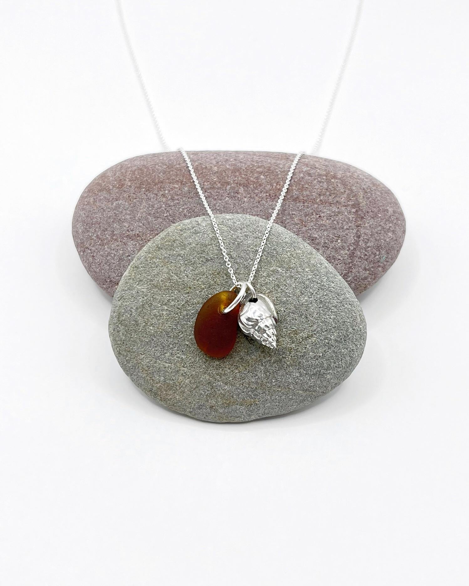 Necklace with burnt orange sea glass and silver shell pendant laid across two pebbles against a white background