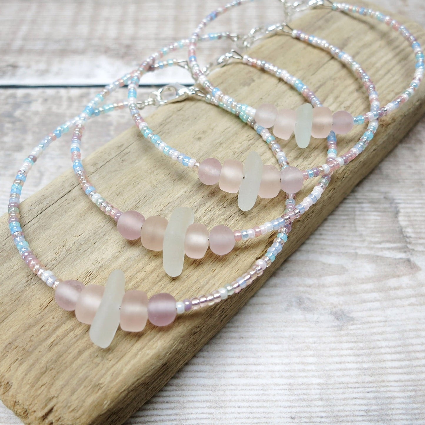 Four bracelets with white sea glass and pastel beads laid on a purple pebble against a wooden background